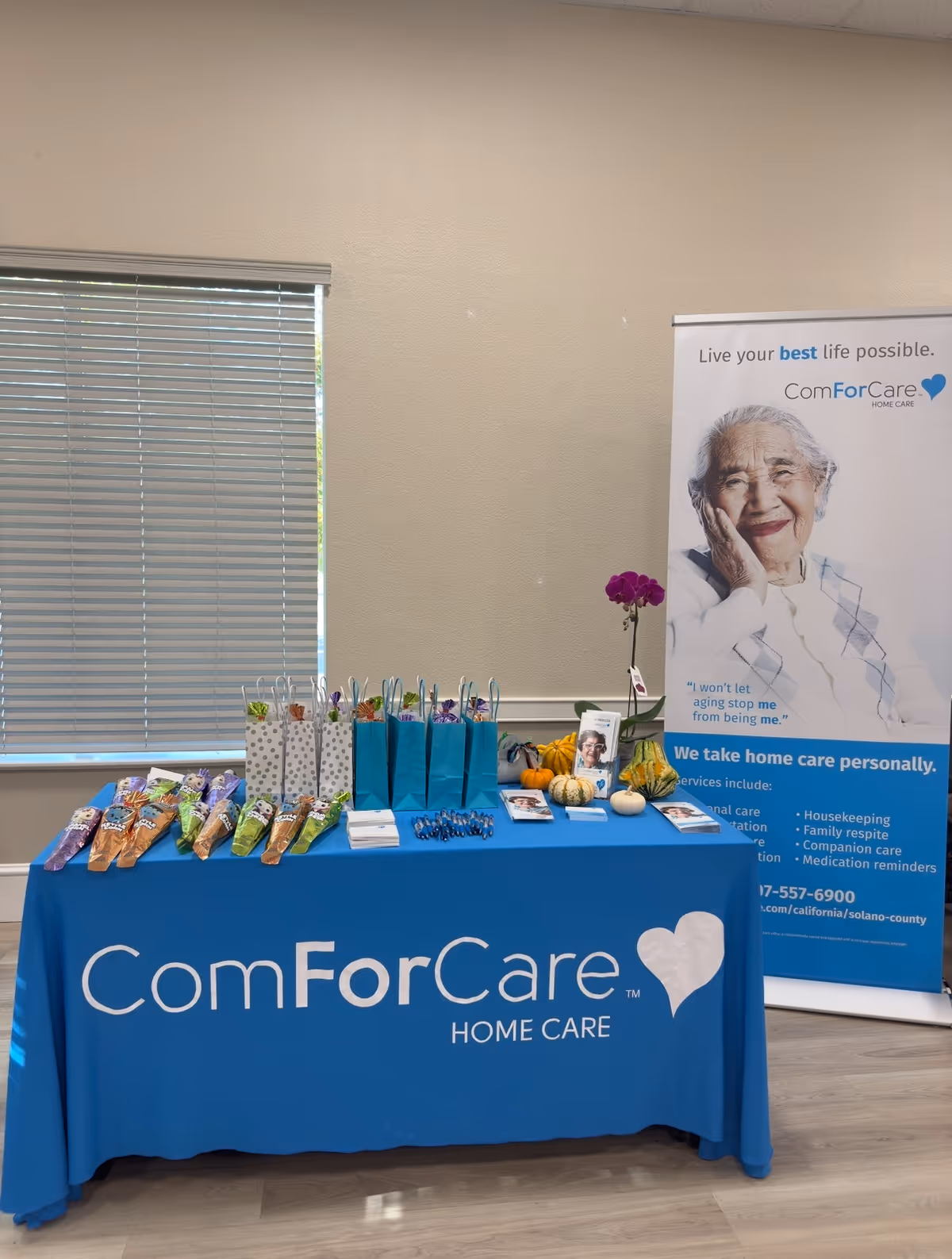 Indoor promotional display table with blue ComForCare branding, gift bags, and a banner.