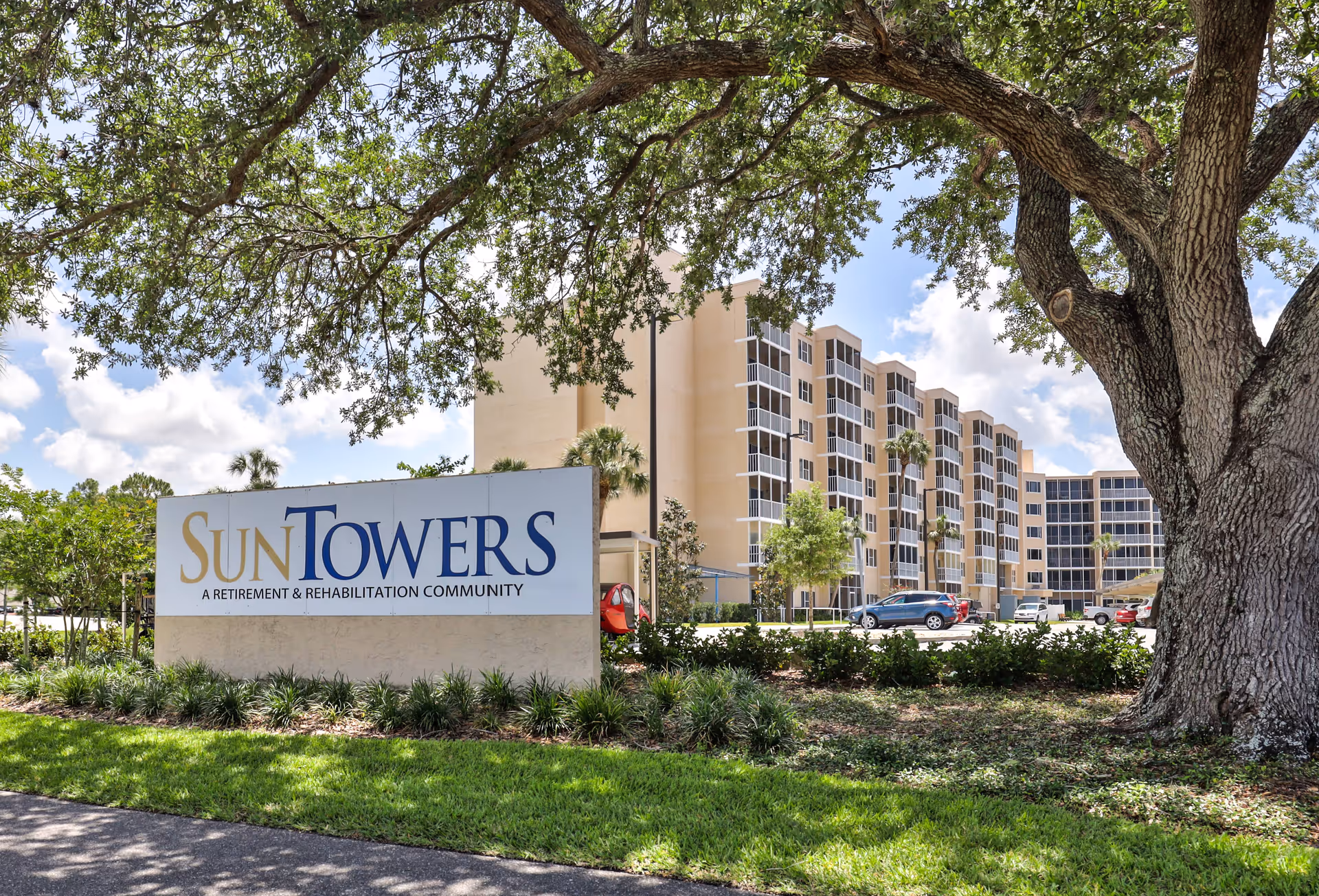 Exterior view of Sun Towers Retirement & Rehabilitation Community building with multiple floors, surrounded by trees and greenery under a partly cloudy sky. A large sign with the community name is visible in the foreground.
