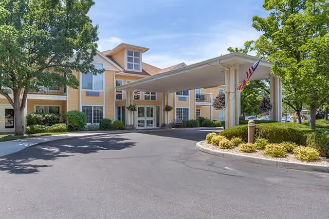 Front exterior view of a senior living facility with a covered entrance, yellow siding, multiple windows, and surrounding trees and landscaping under a clear blue sky.
