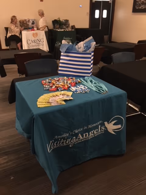 Promotional table with Visiting Angels logo, brochures, and candy in a conference room.