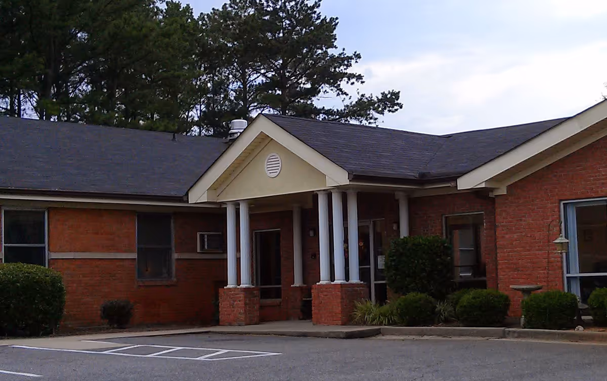 Front entrance of a single-story brick senior living building with white columns and a small parking area.