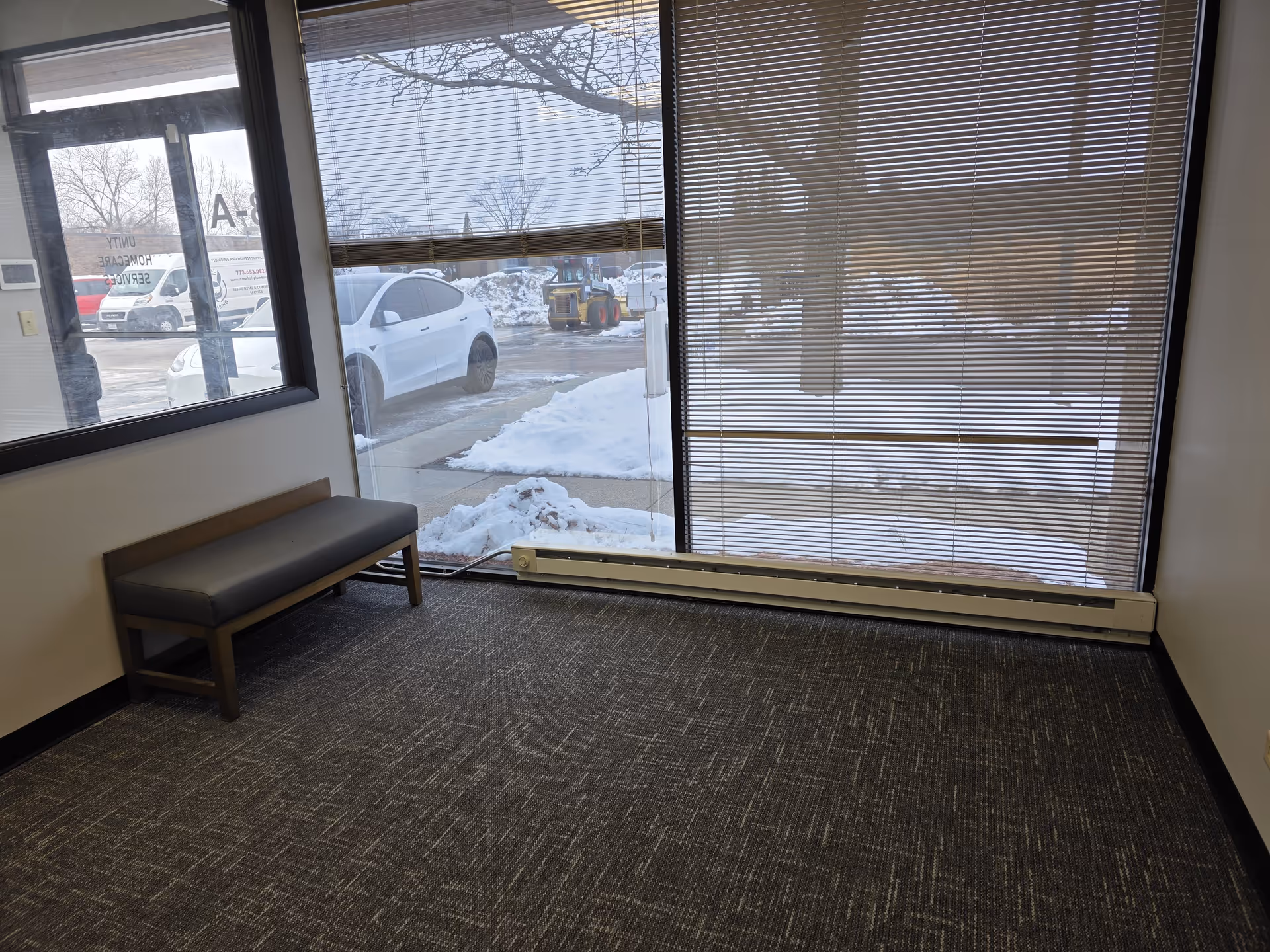 A small waiting area with a bench by large windows and blinds in a winter scene.