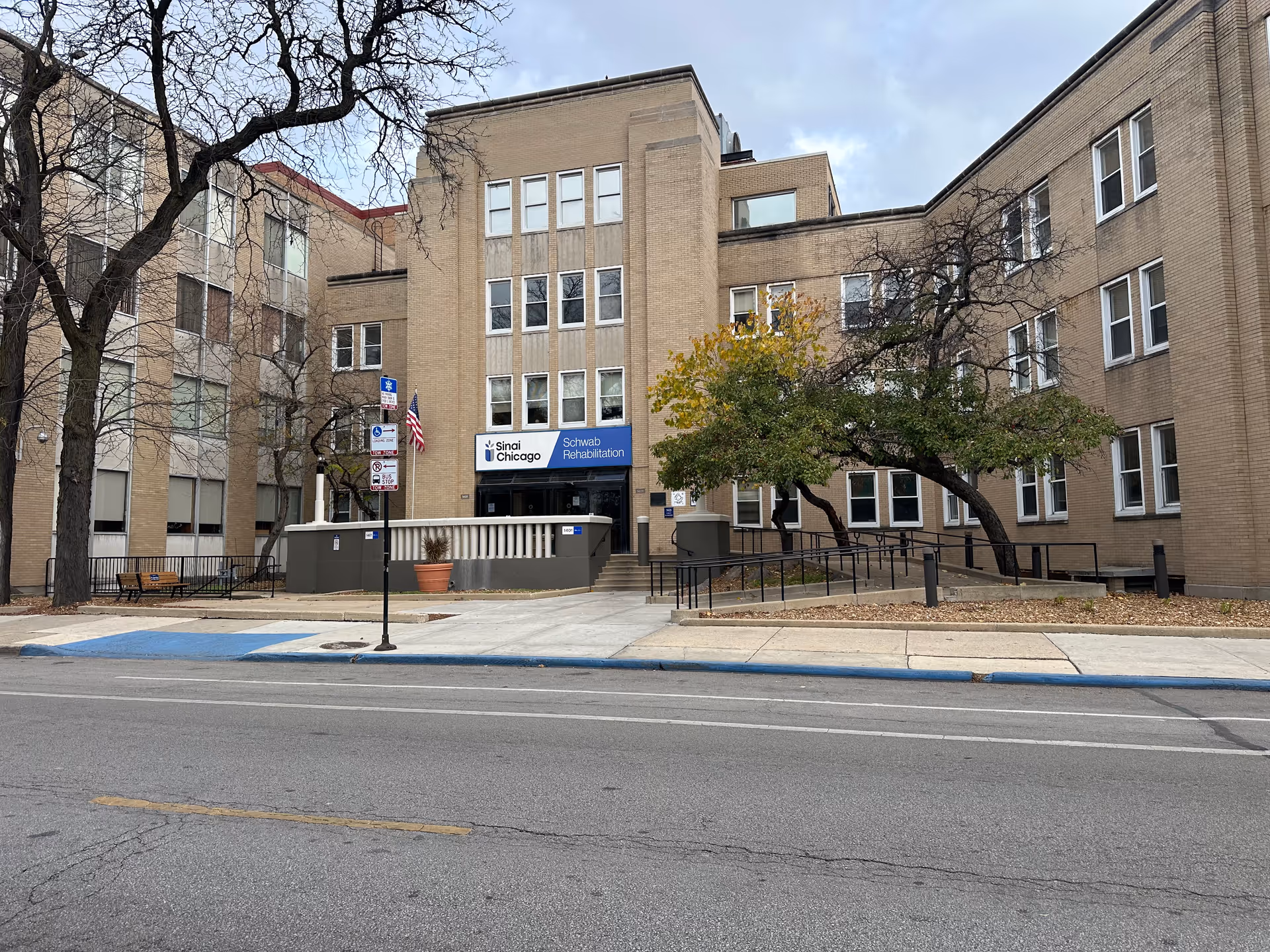 Front exterior of a multi-story brick rehabilitation center (Sinai Chicago Schwab Rehabilitation) with an entrance, ramp, flag, and trees.