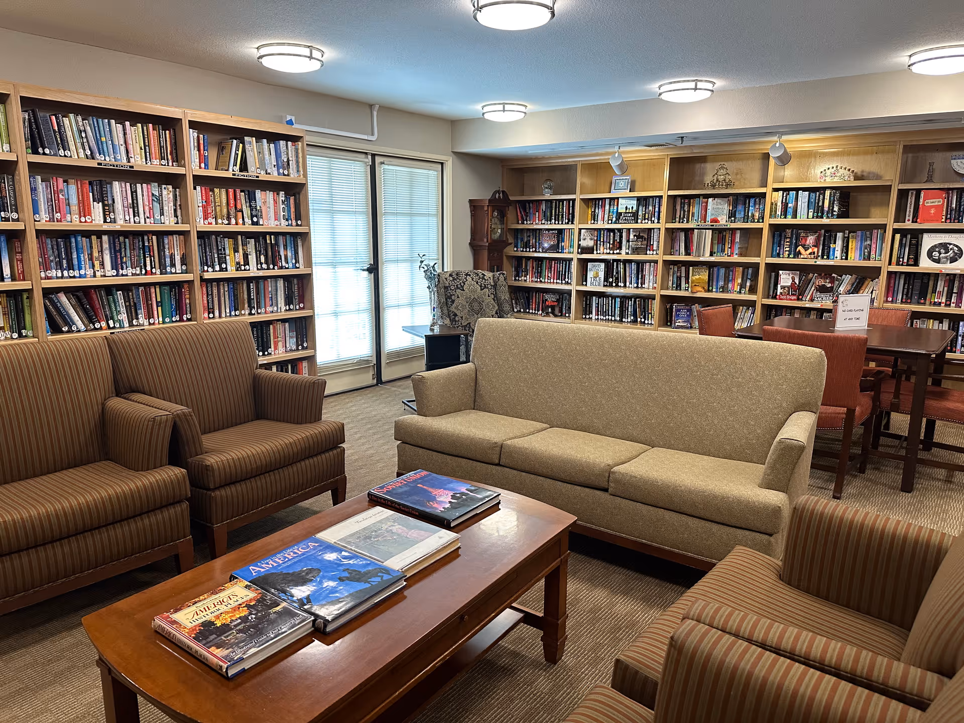 A cozy library room with multiple bookshelves filled with books along the walls. The room features a beige three-seat sofa, two striped armchairs, and a wooden coffee table with several large books on it. There is a window with blinds and a door letting in natural light, and a wooden table with chairs in the background.