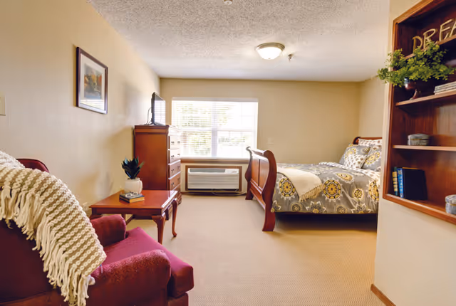 Furnished bedroom with a wooden sleigh bed, dresser and TV, armchair, side table, and a window air conditioner.