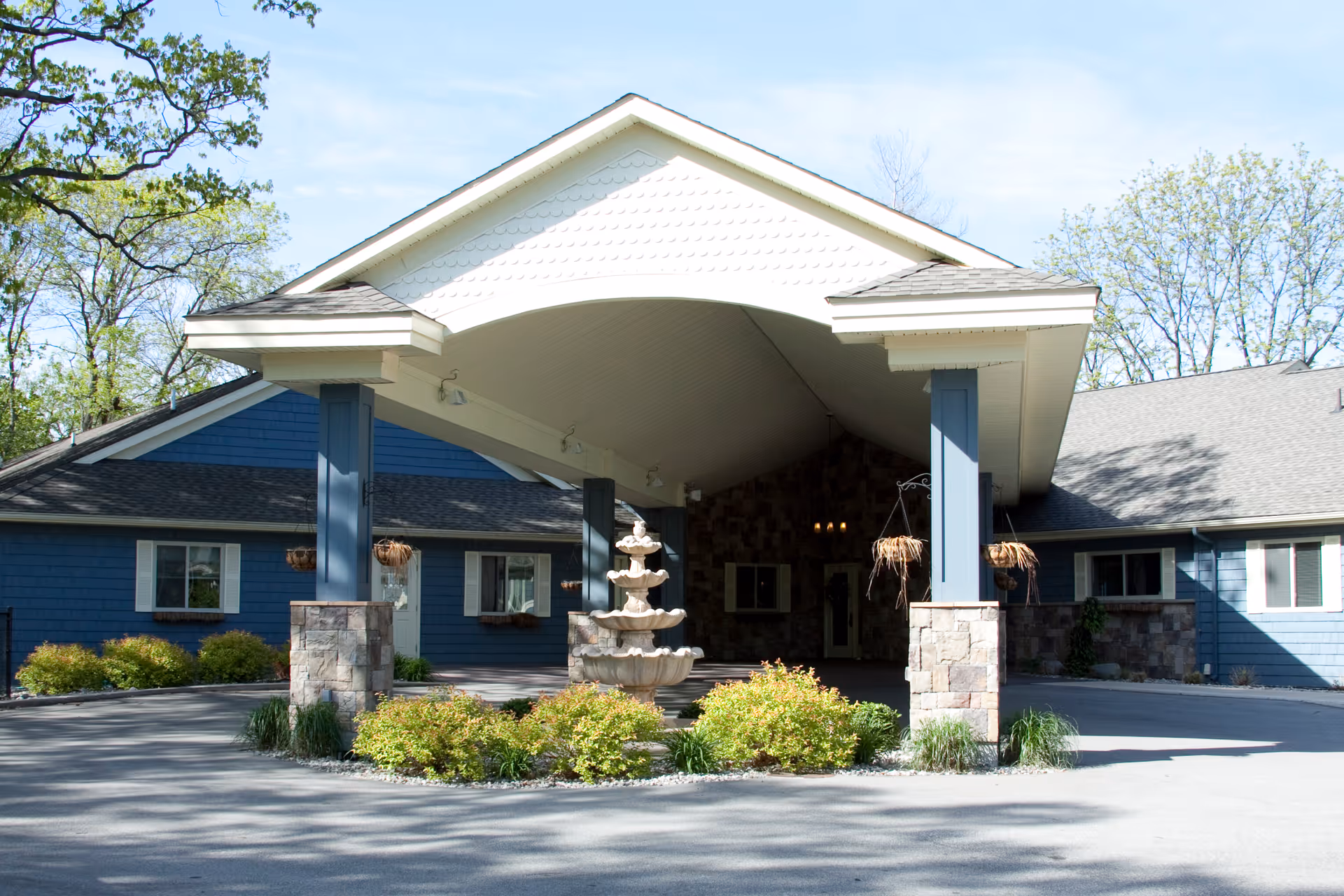 Front exterior view of Stone Crest Assisted Living facility featuring a covered entrance with stone pillars, a multi-tiered fountain surrounded by greenery, and blue siding on the building under a clear sky.