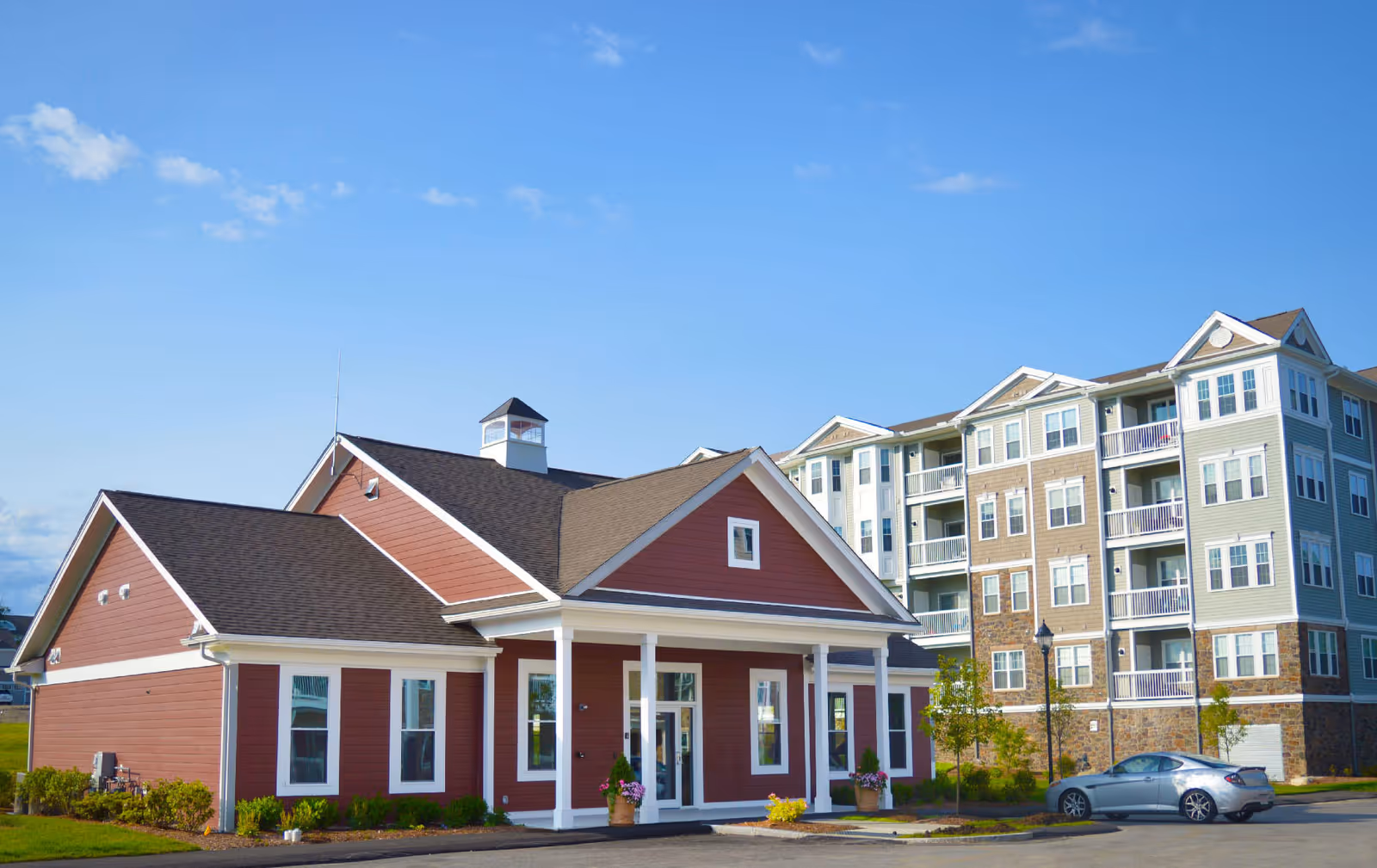 Exterior view of a senior living facility with a red building featuring white trim and a covered entrance in the foreground, and a larger multi-story residential building with balconies in the background under a clear blue sky.