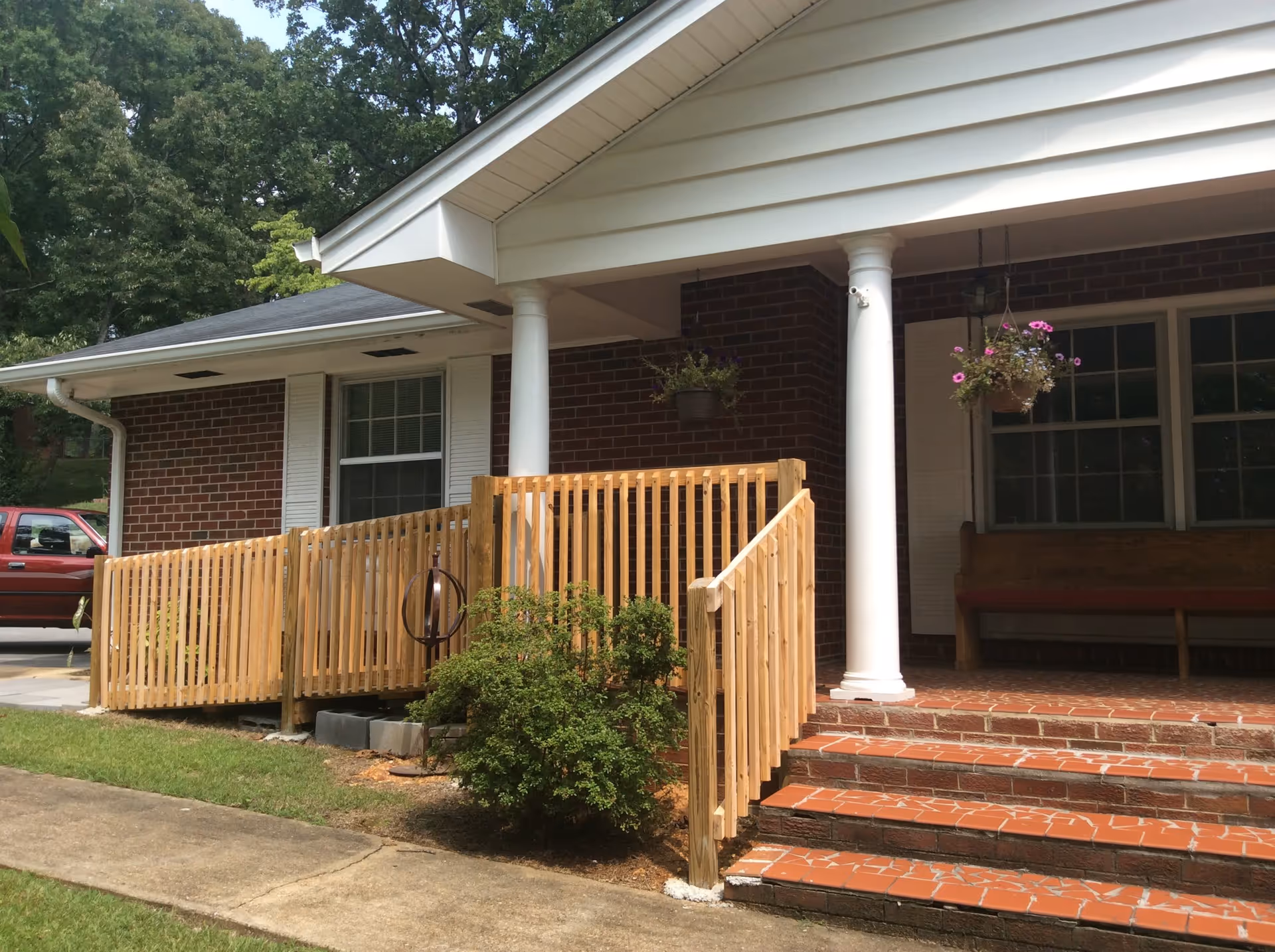 Exterior view of a brick building with white columns and a wooden ramp leading to the entrance. There are hanging flower pots with pink flowers and a wooden bench on the porch. A red vehicle is partially visible on the left side, and there are trees in the background.