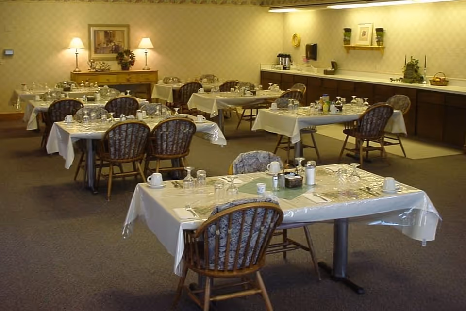 A dining room set up with multiple tables covered in white tablecloths and plastic covers, each table set with cups, glasses, and silverware. The room has patterned wallpaper, carpeted floor, wooden chairs with cushions, and a sideboard with lamps and decorative items.