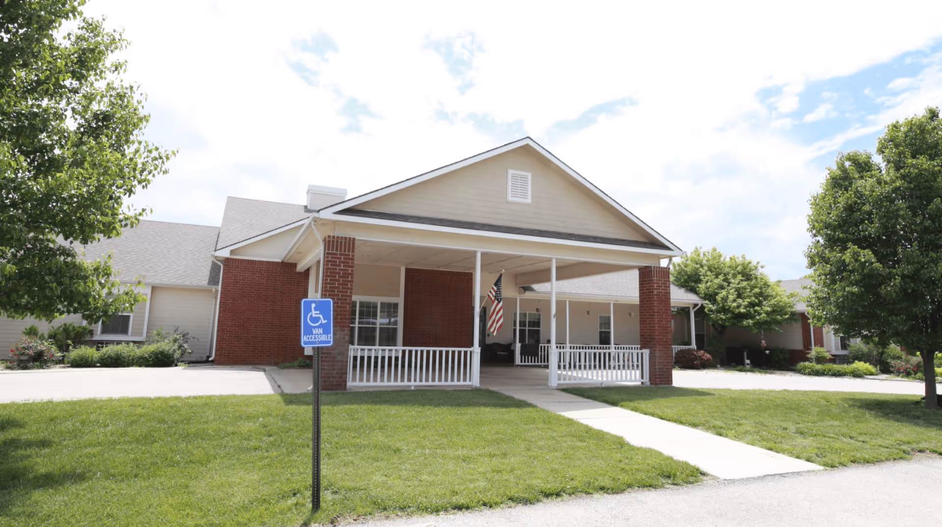 Front entrance of a single-story assisted living building with a covered drop-off, American flag, and a handicap parking sign on a grassy lawn.