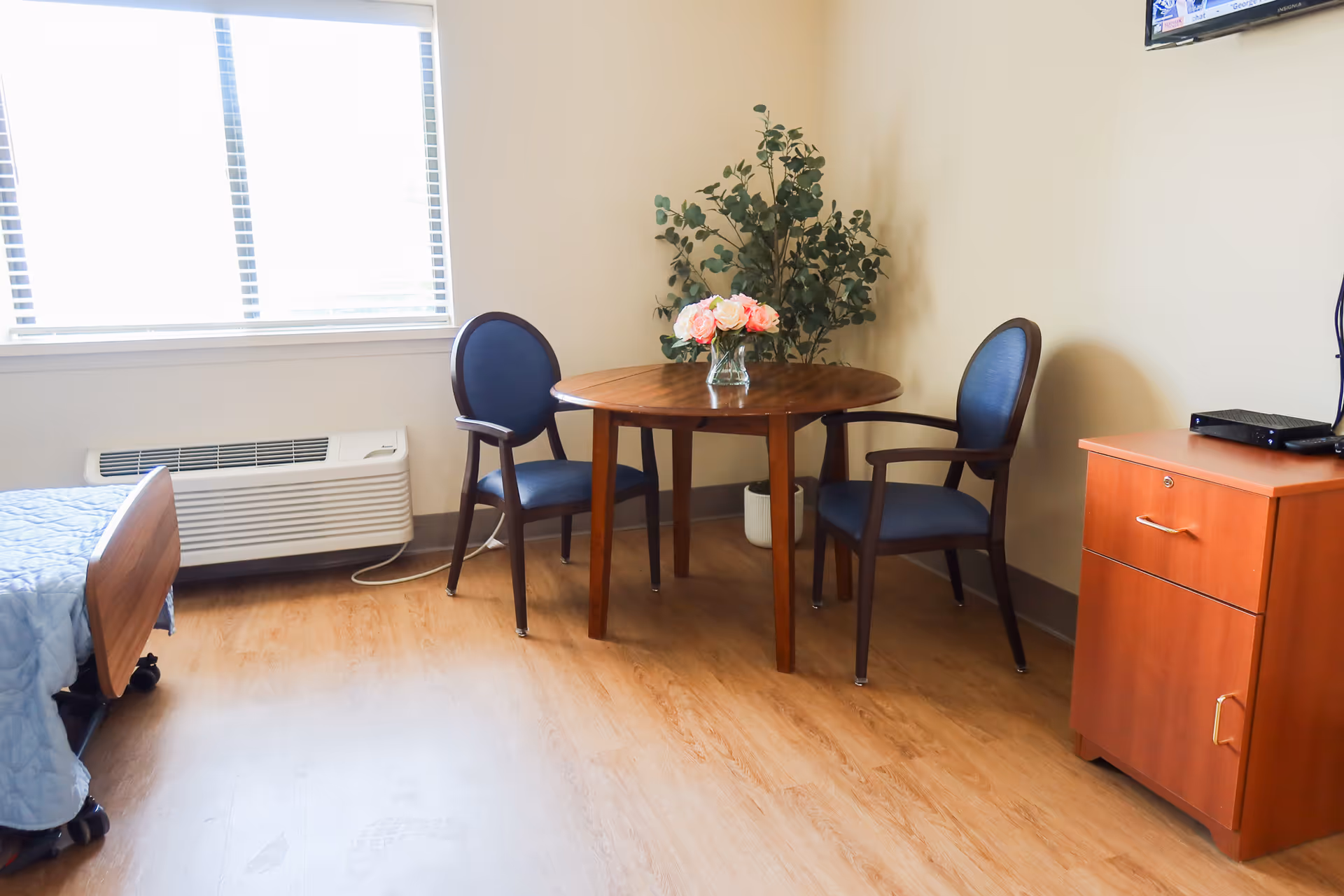 A small furnished hospital-like bedroom with a wooden dining table, three blue chairs, and a plant.