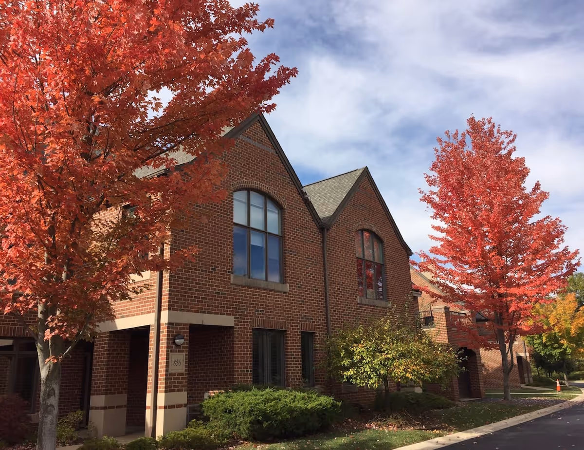 Brick two-story building front with arched windows and bright red autumn trees along a driveway.