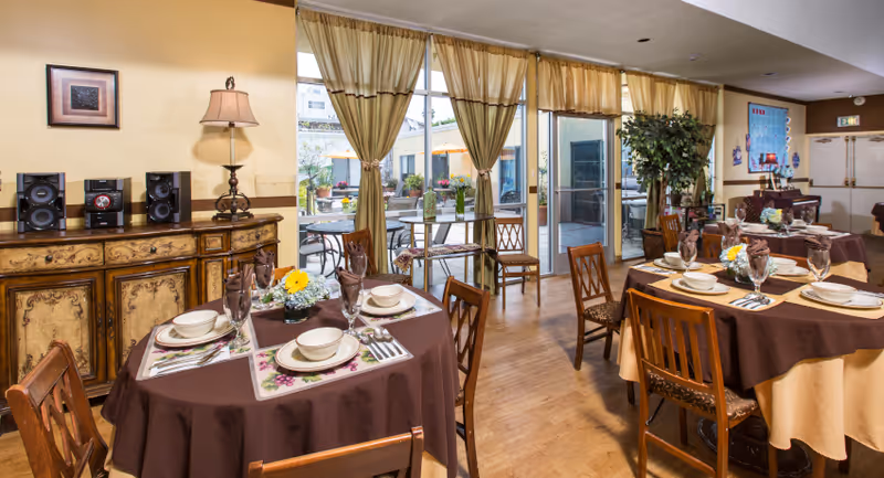 Dining room with round tables set for meals, wooden chairs, and large windows with drapes opening to a patio.