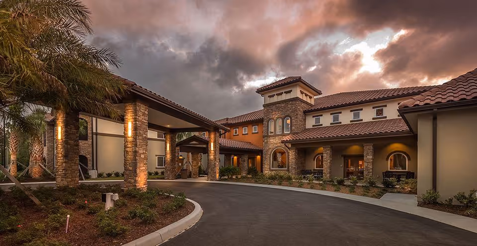 Entrance and porte-cochère of a Mediterranean-style senior living building at dusk with illuminated stone columns and a curved driveway.