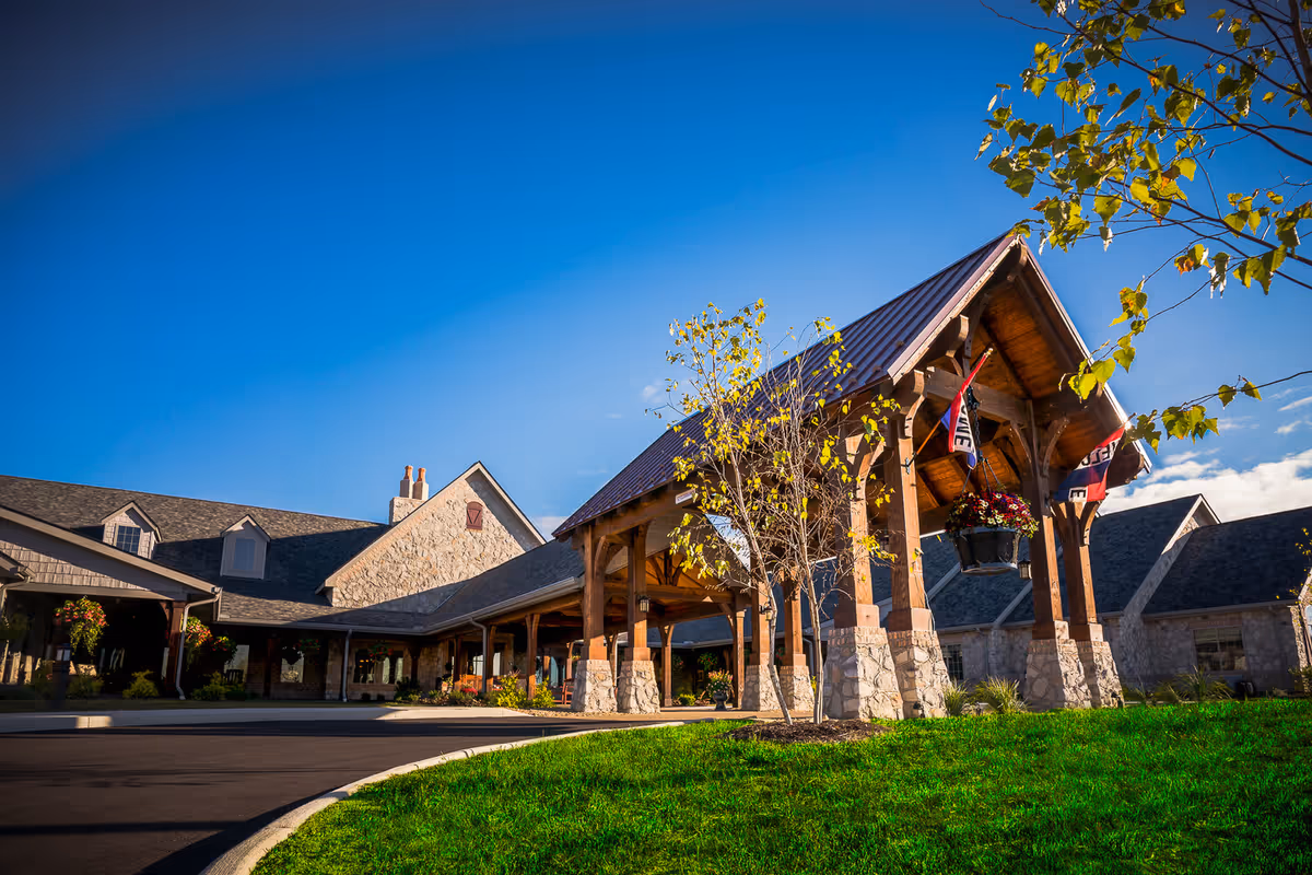 Exterior view of The Inn at Walnut Trail showing a large stone building with a covered entrance supported by wooden beams, surrounded by green grass and small trees under a clear blue sky.