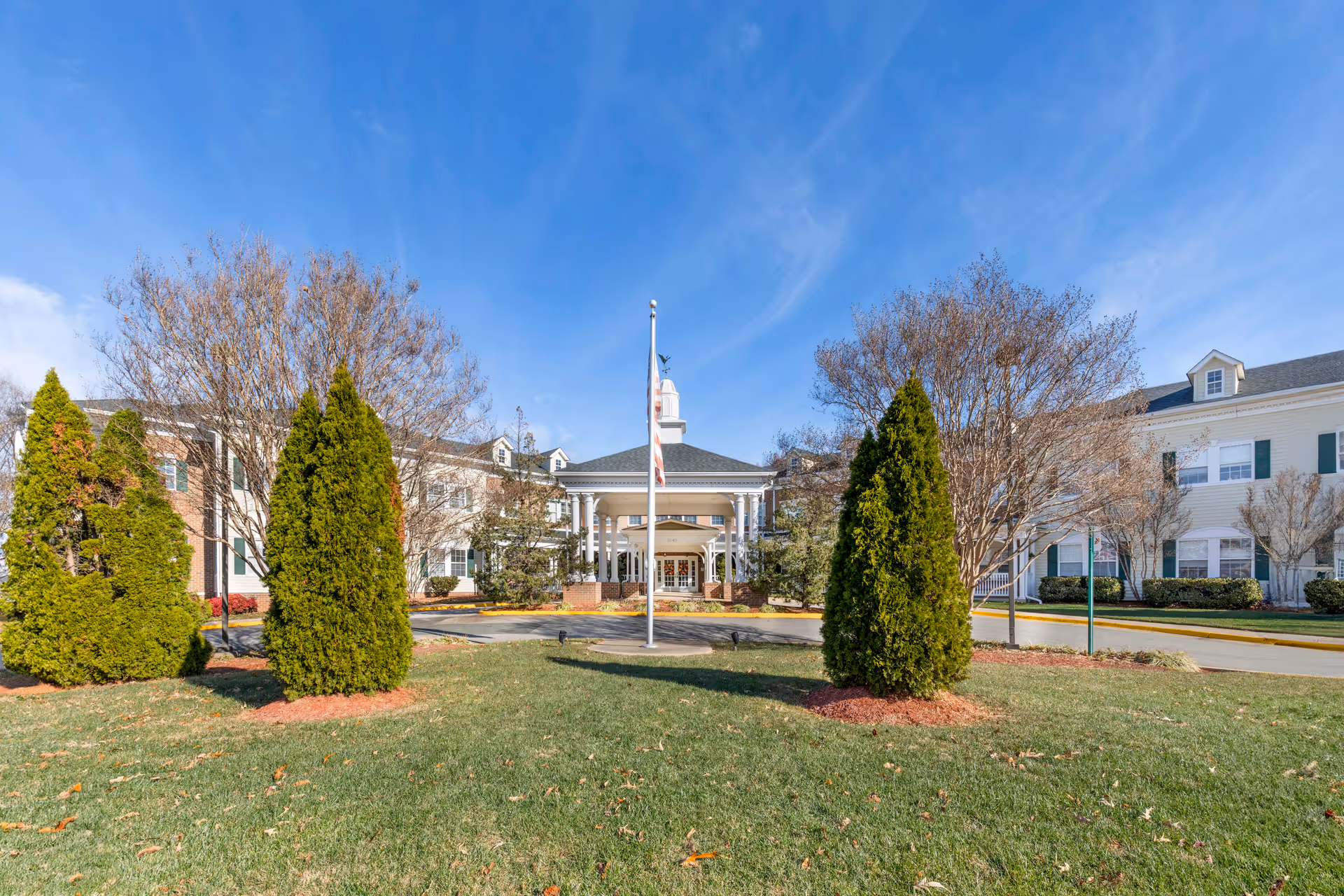 Brookdale Lake Ridge front entrance with flagpole, manicured landscaping, and a blue sky.