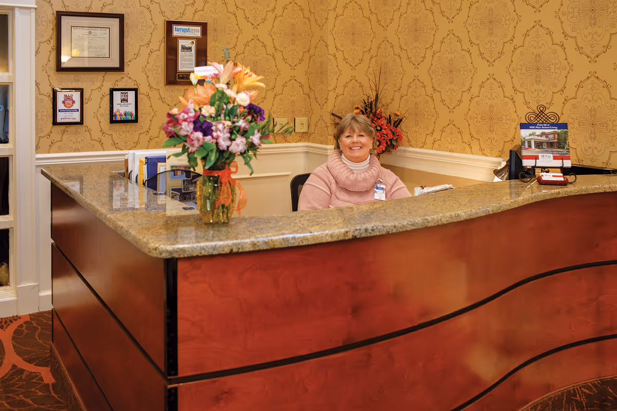 Reception desk inside a senior living facility with a smiling receptionist seated behind a curved wood counter and a vase of flowers on the countertop.