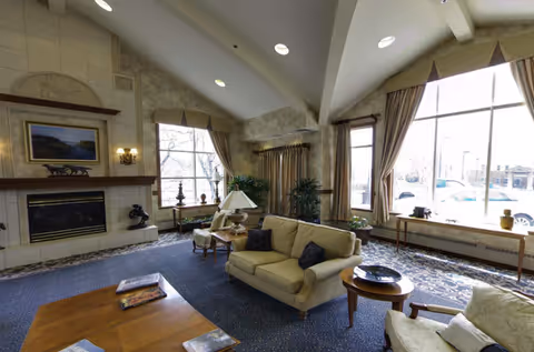 A spacious living room with high vaulted ceilings, large windows with curtains letting in natural light, a fireplace with a painting above it, beige sofas and armchairs arranged around a coffee table, and decorative plants near the windows.