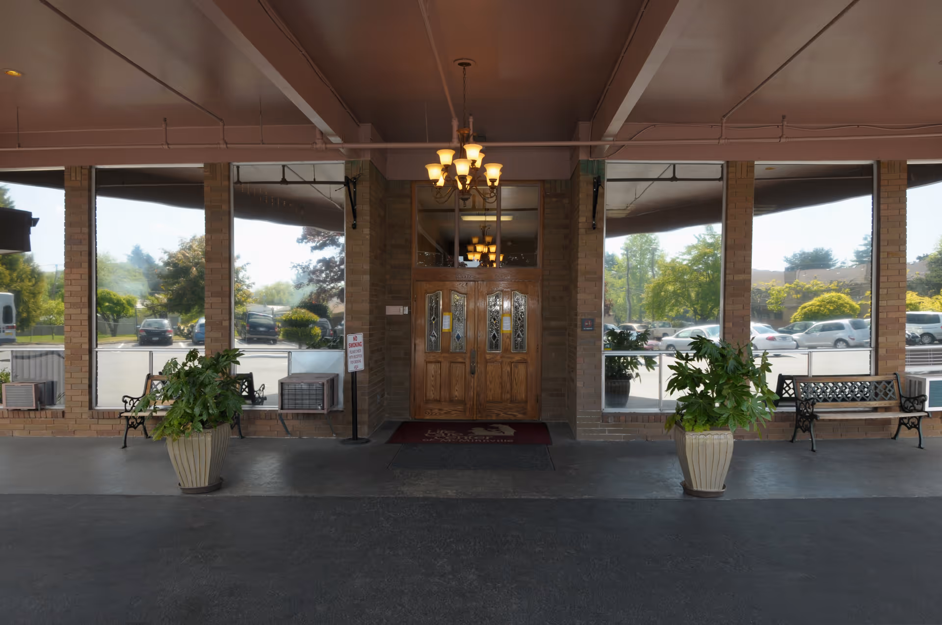 Entrance of a building with double wooden doors featuring glass panels, flanked by large windows. Two potted plants are placed symmetrically on either side of the entrance, and two metal benches are positioned near the windows. A chandelier with multiple lights hangs from the ceiling above the entrance. Outside, cars and greenery are visible through the windows.