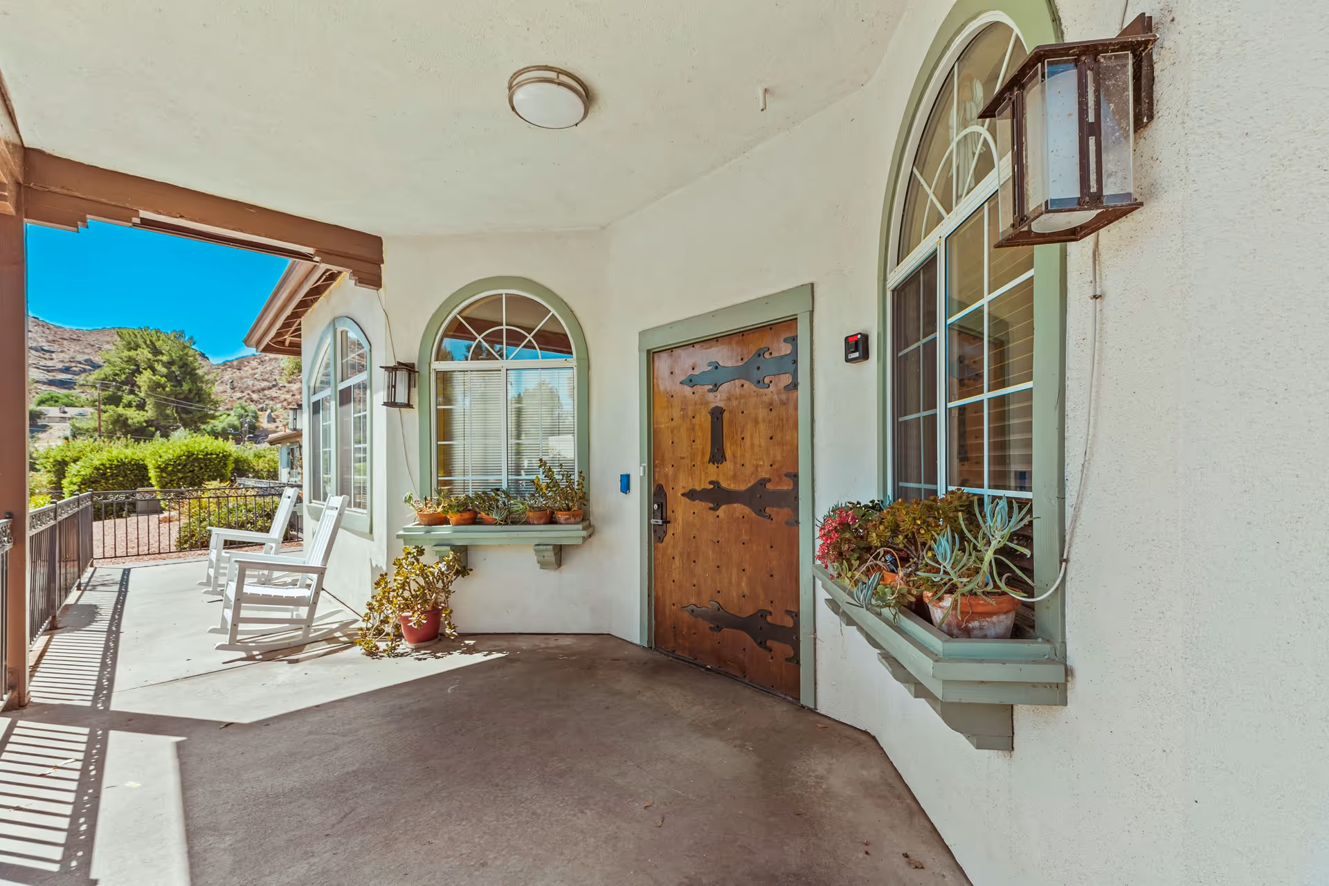 Covered porch area with a wooden door featuring decorative black hinges, two arched windows with green trim and flower pots on the window sills, two white rocking chairs, and outdoor lantern-style light fixtures. The porch overlooks a garden with bushes and trees under a clear blue sky.