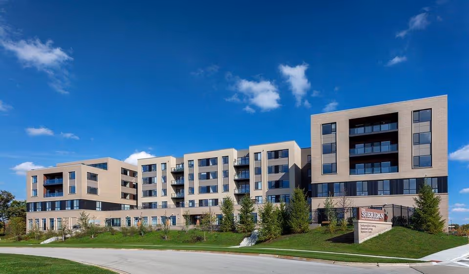 Modern multi-story beige senior living building with balconies, landscaped lawn, and a sign under a blue sky.