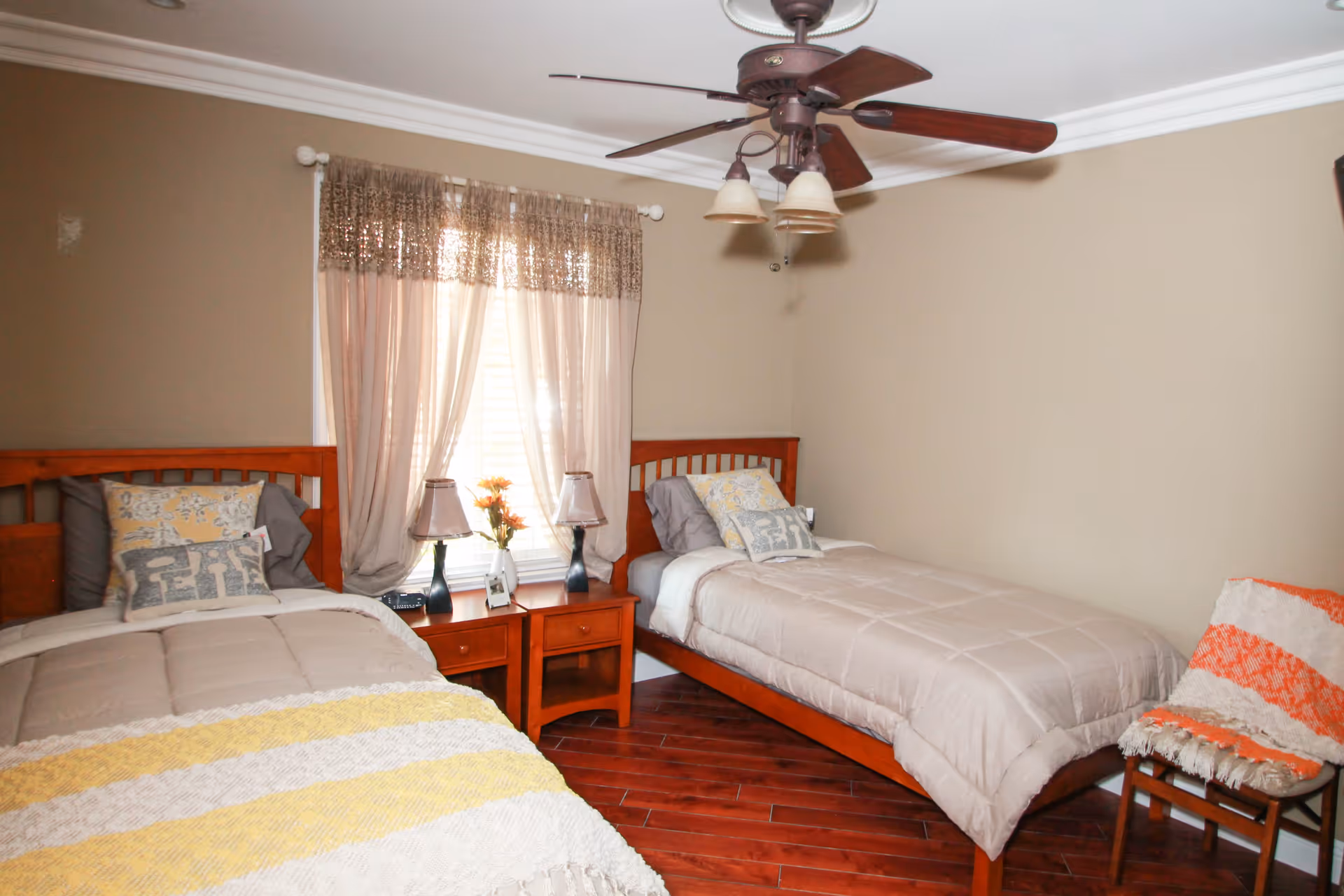 A bedroom with two single wooden beds, each with beige bedding and decorative pillows. Between the beds is a wooden nightstand with two lamps and a small flower arrangement. A window with sheer curtains is behind the nightstand. There is a wooden chair with an orange and white striped blanket on the right side of the room. The floor is wooden, and a ceiling fan with lights is mounted on the ceiling.
