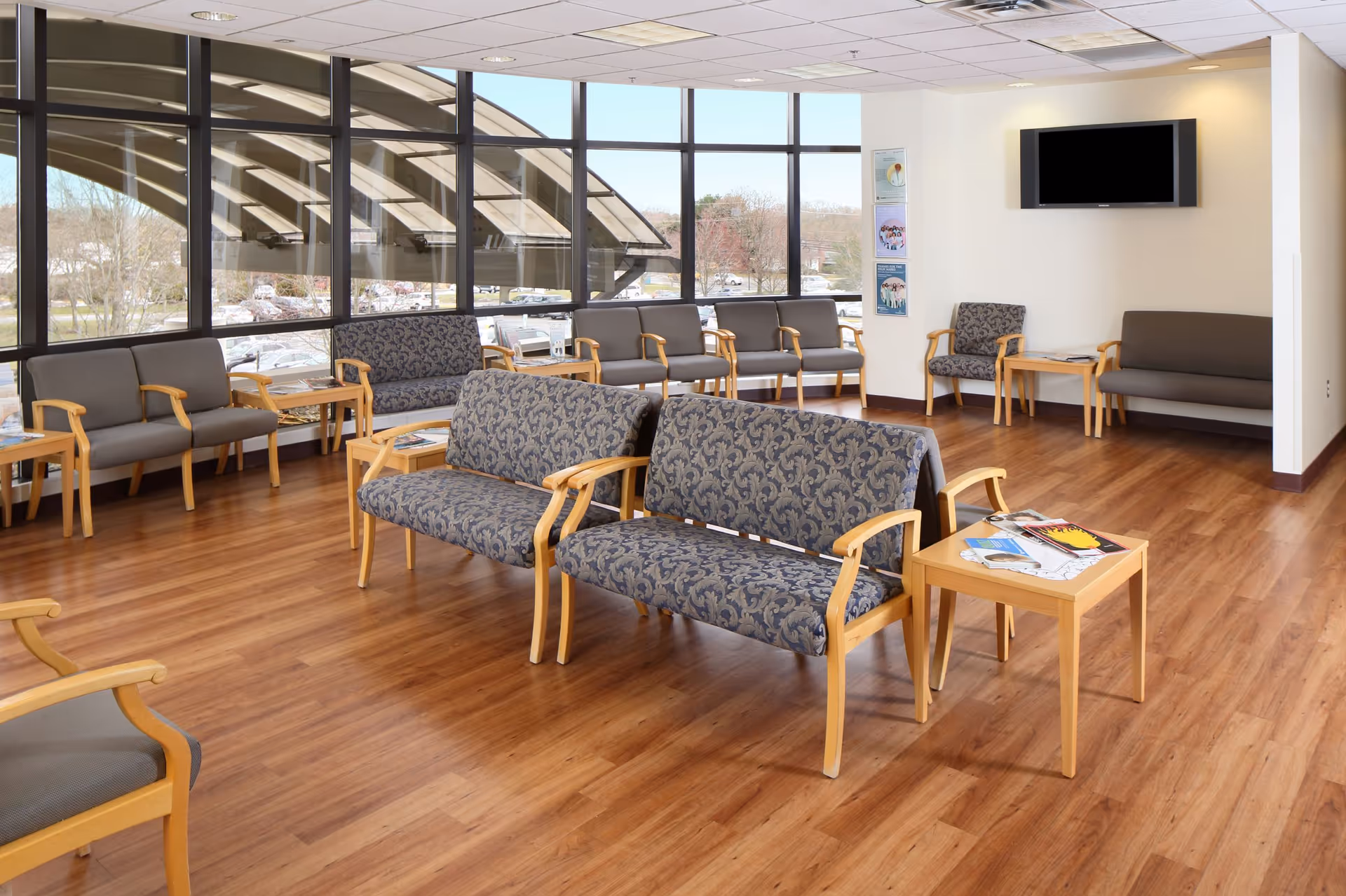 A spacious waiting lounge with multiple sofas, chairs, magazines, and a wall-mounted TV by large windows.