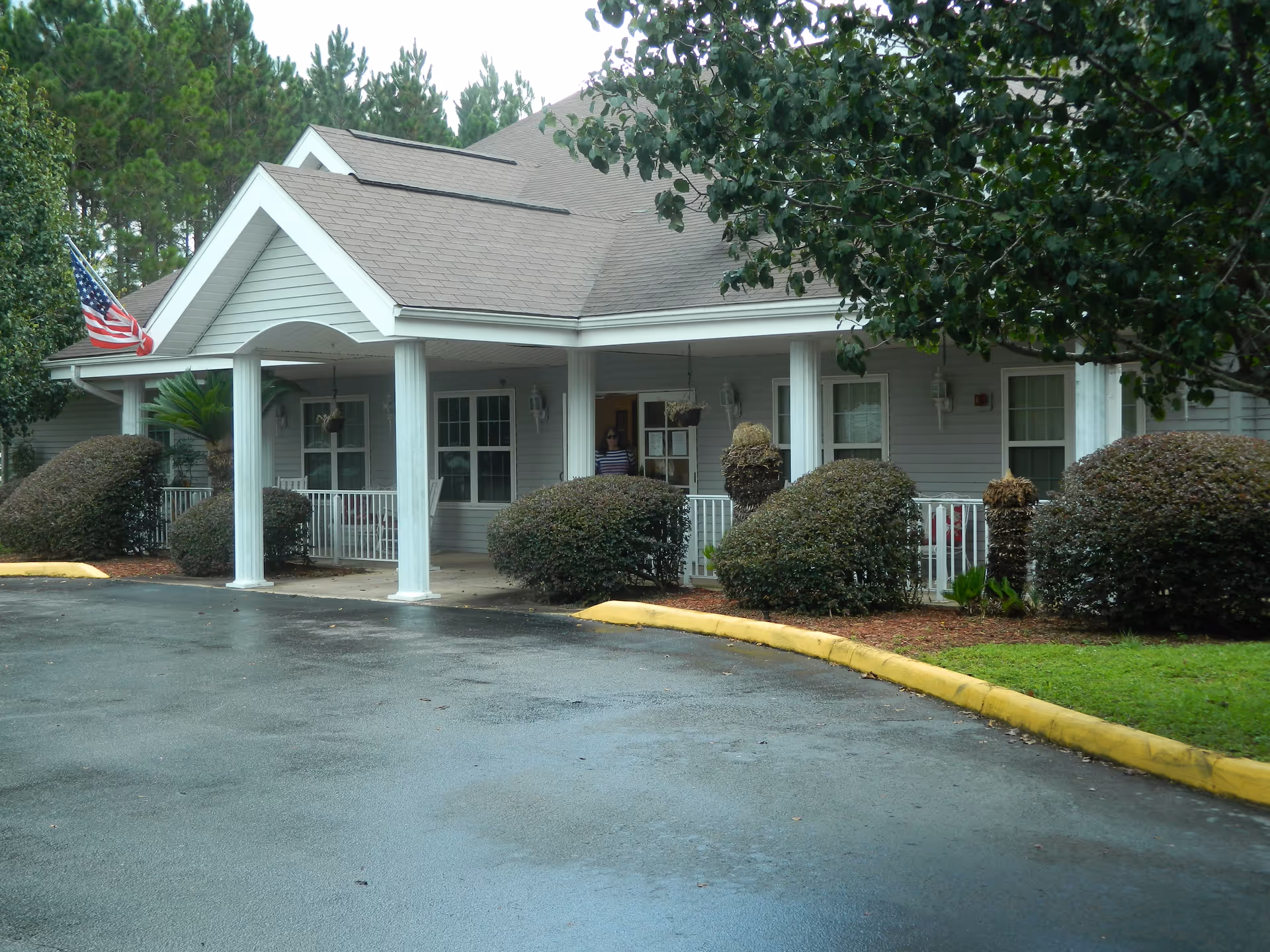 Single-story senior living facility with a covered front entrance, pillars, flag, and shrubs.