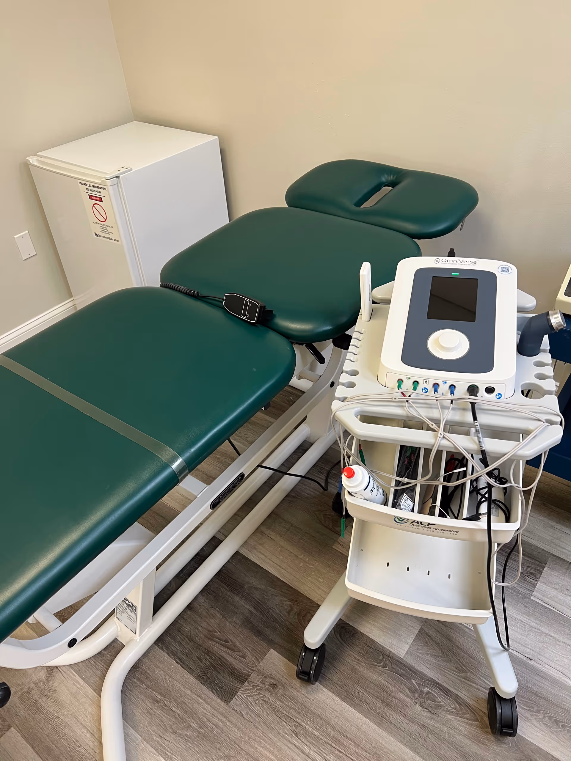 Green padded treatment table with a medical cart and monitor equipment in a clinical therapy room.