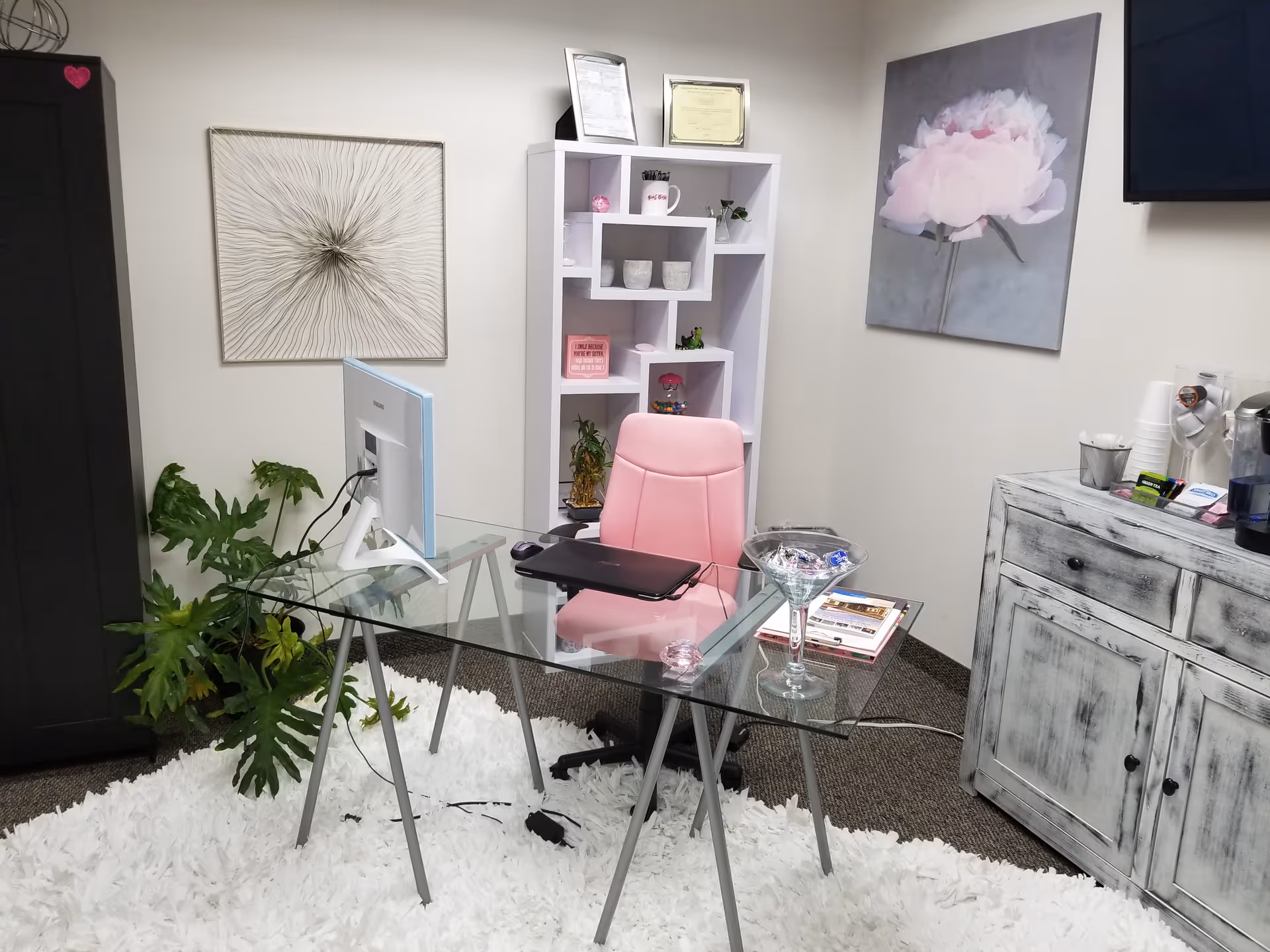 A glass desk with a pink chair, white rug, and decorative shelving in an office.