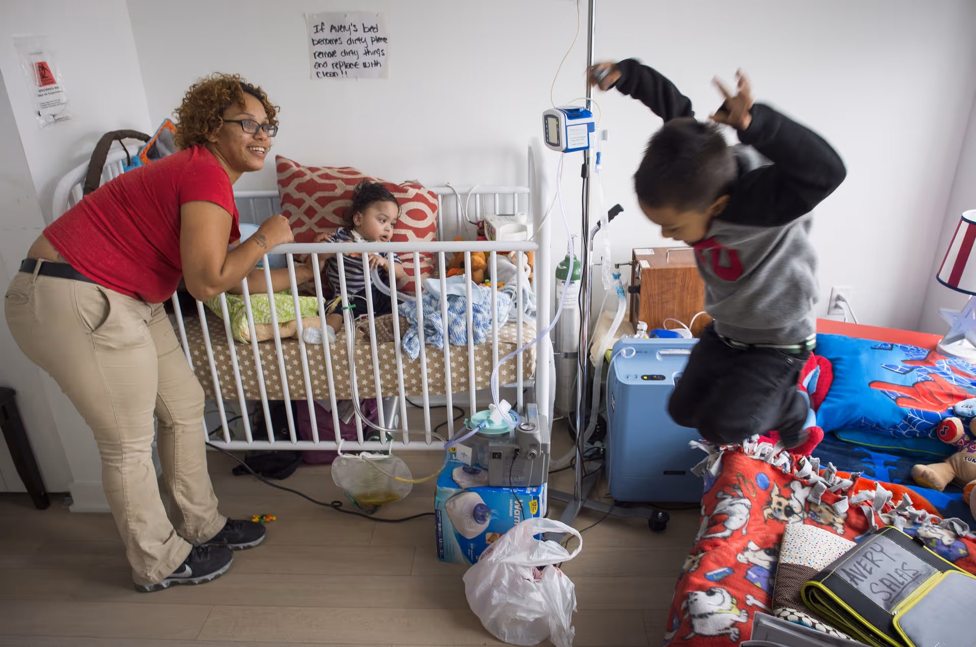 A caregiver leans by a hospital bed while two children play in a medical care room.