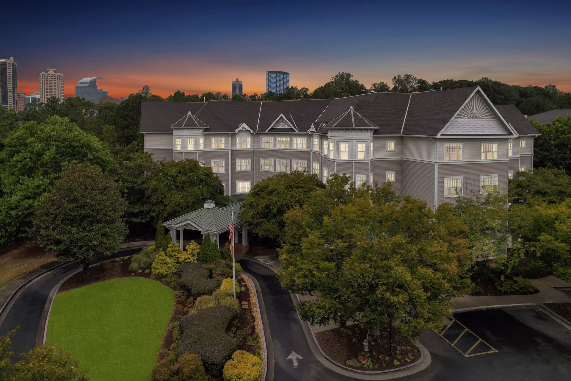 Exterior view of a large senior living facility building named Sunrise at Buckhead during sunset, surrounded by trees and landscaped greenery with a driveway and a small gazebo at the entrance.