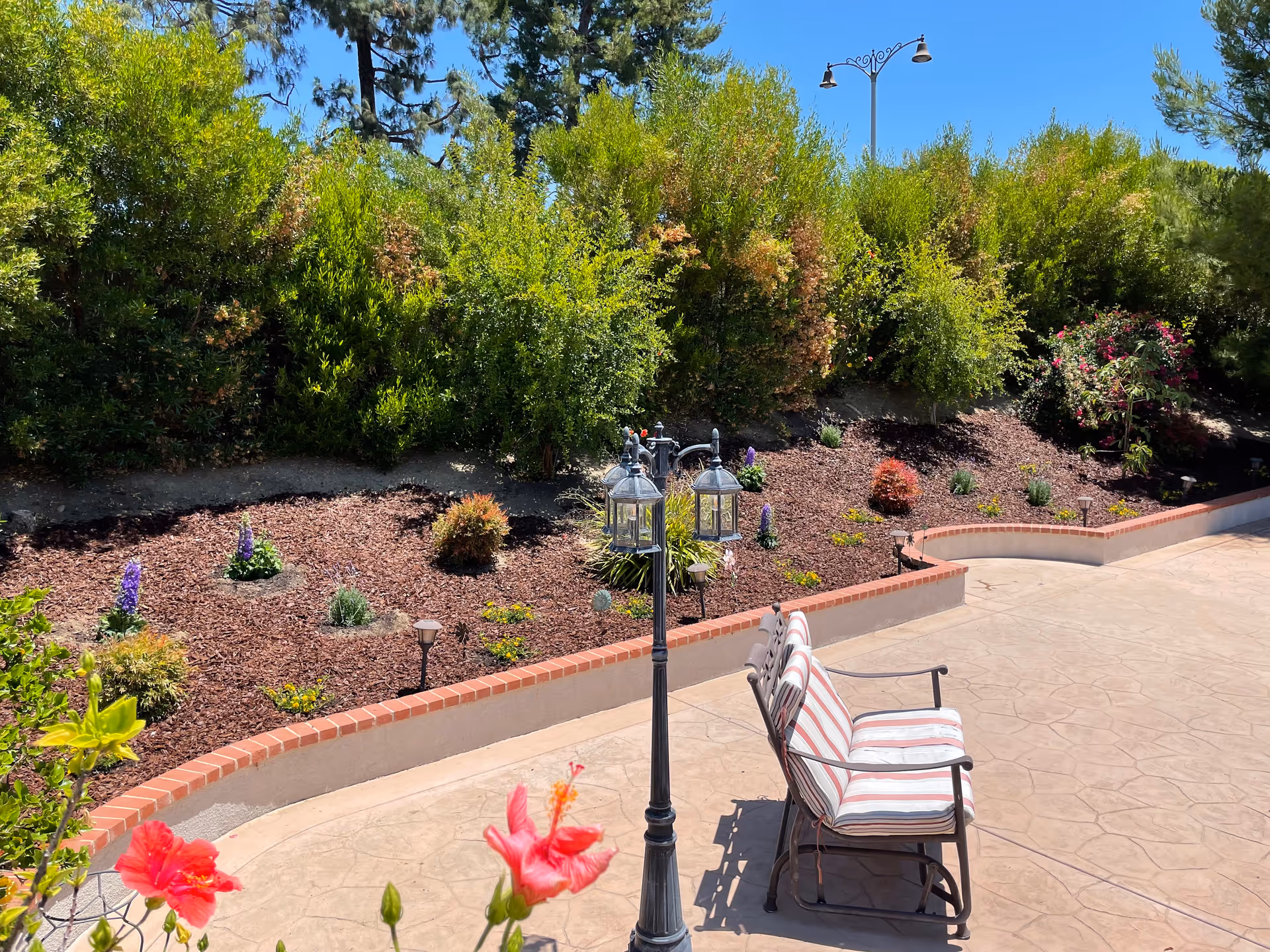 Sunny outdoor patio with a striped bench, decorative lamppost, red flowers and landscaped planting beds.