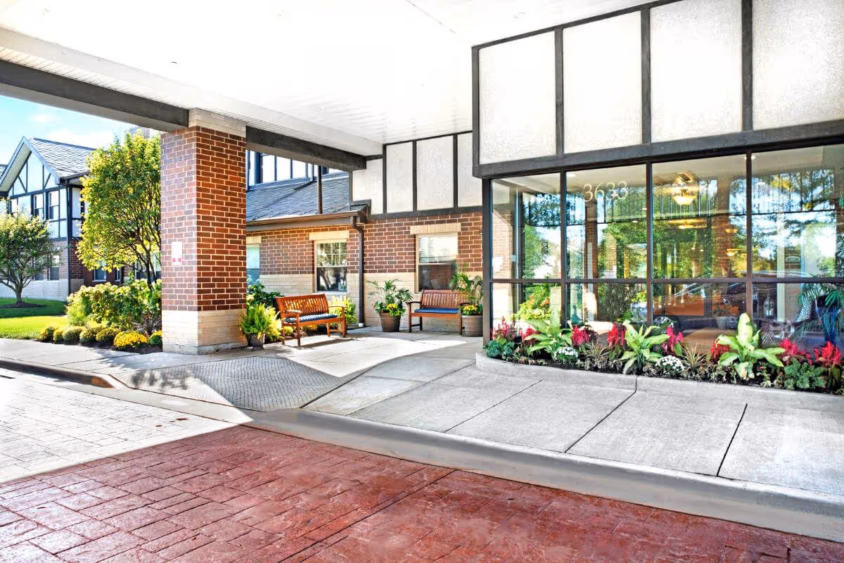 Covered front entrance of a senior living facility with benches, potted plants, flower beds and large glass doors.
