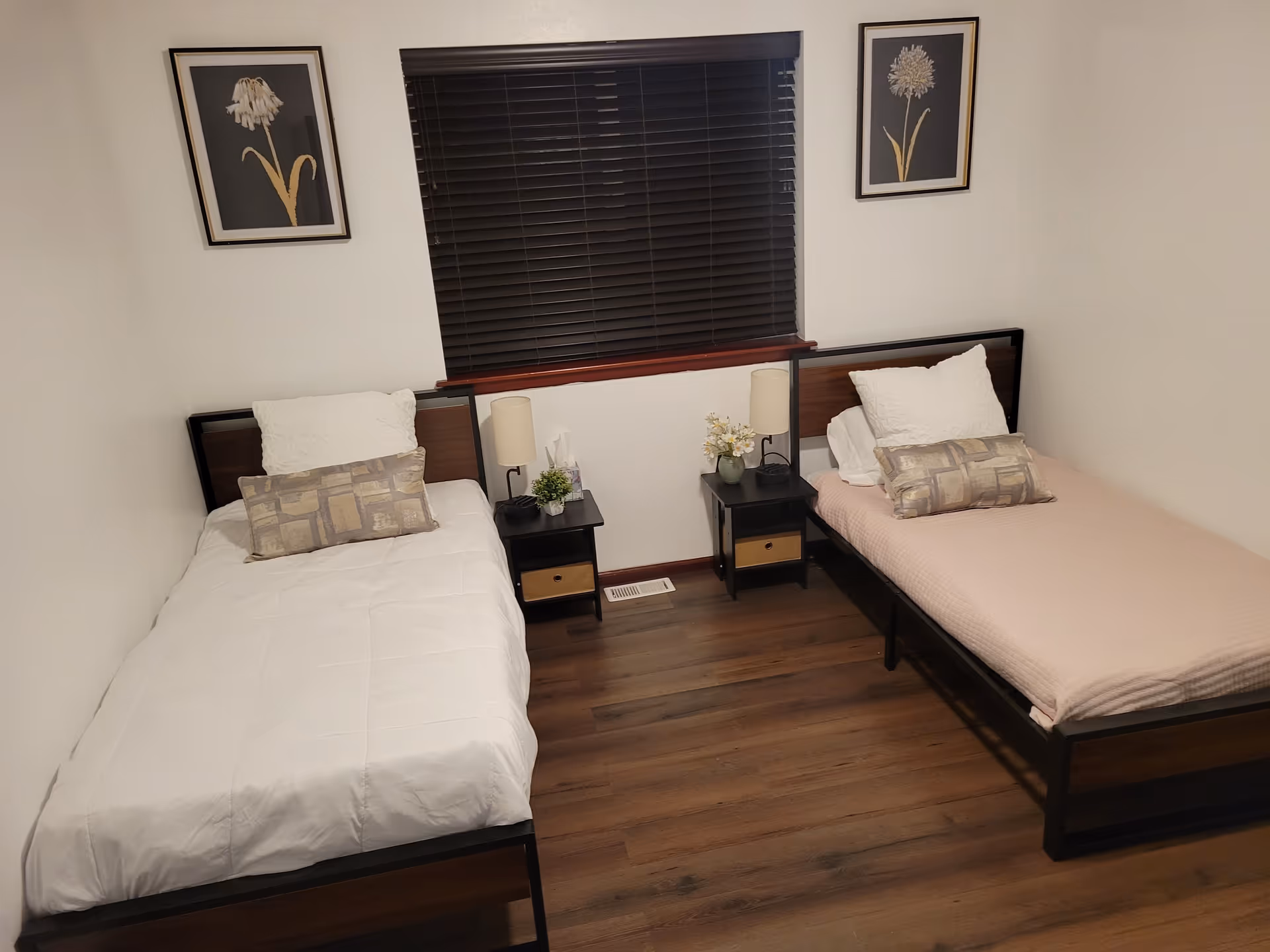 Two single beds, nightstands, window with blinds, and floral wall art in bedroom.