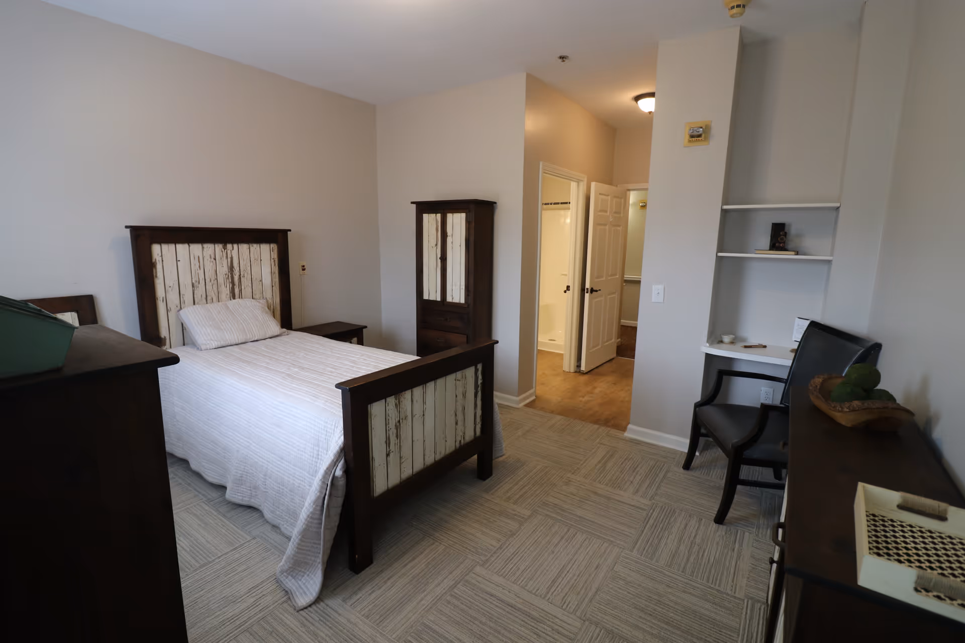 A furnished memory care bedroom with a bed, dresser, wardrobe, and seating area.