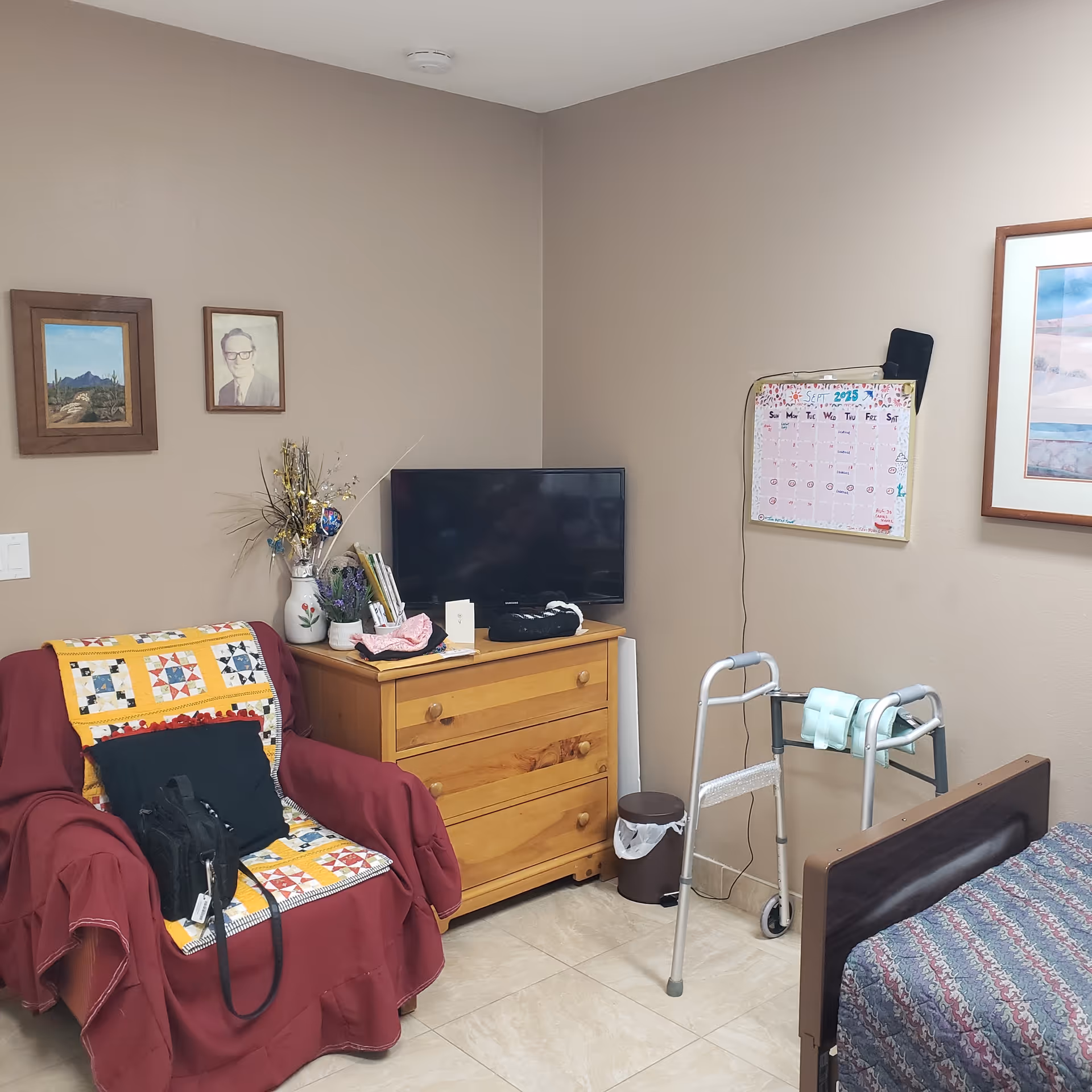 Cozy resident room featuring a bed, walker, dresser with TV, and quilted chair nearby.