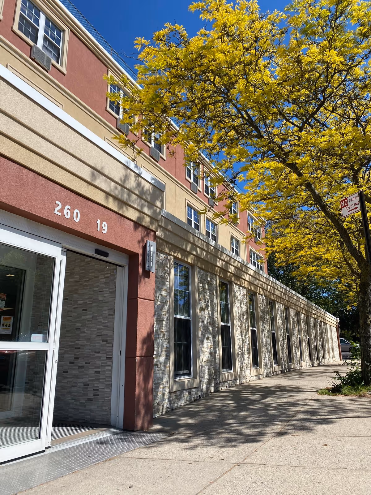 Exterior view of a multi-story building with beige and red walls, large windows, and an entrance with sliding glass doors. A tree with yellow flowers is on the right side casting shadows on the sidewalk and building. The sky is clear and blue.