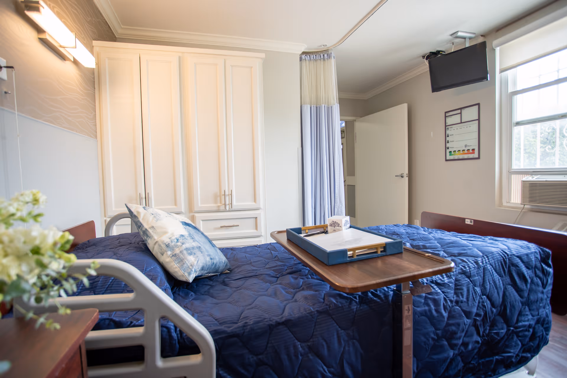 A bright and clean senior living bedroom with a hospital-style bed covered in a dark blue quilt and a decorative pillow. A wooden overbed table with a tray and tissue box is positioned over the bed. The room features white cabinetry, a wall-mounted TV, a window with an air conditioning unit, and a privacy curtain near the door. A small vase with flowers is visible on a bedside table.