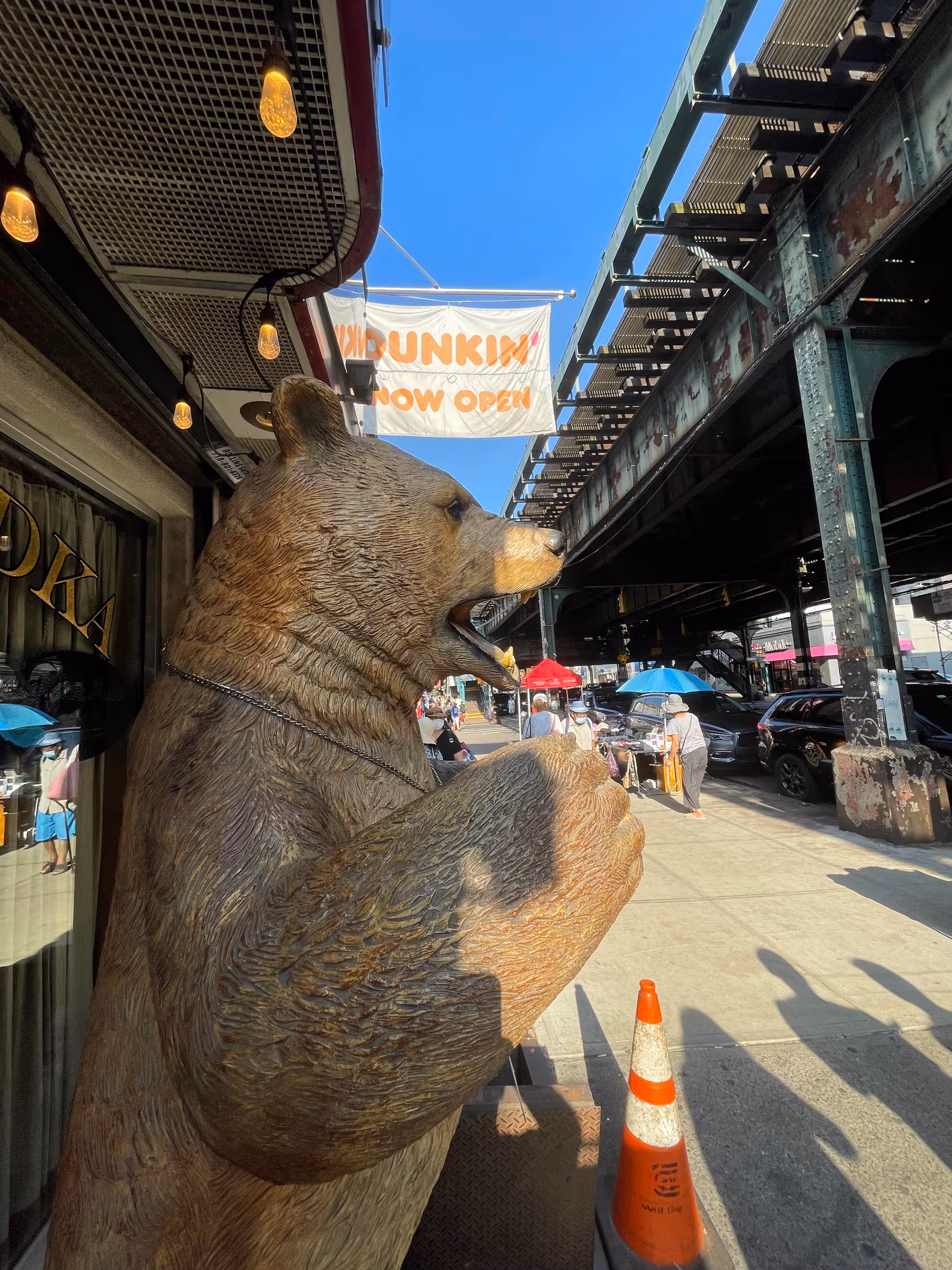 Large bear statue on a city sidewalk beside a Dunkin banner and pedestrians.
