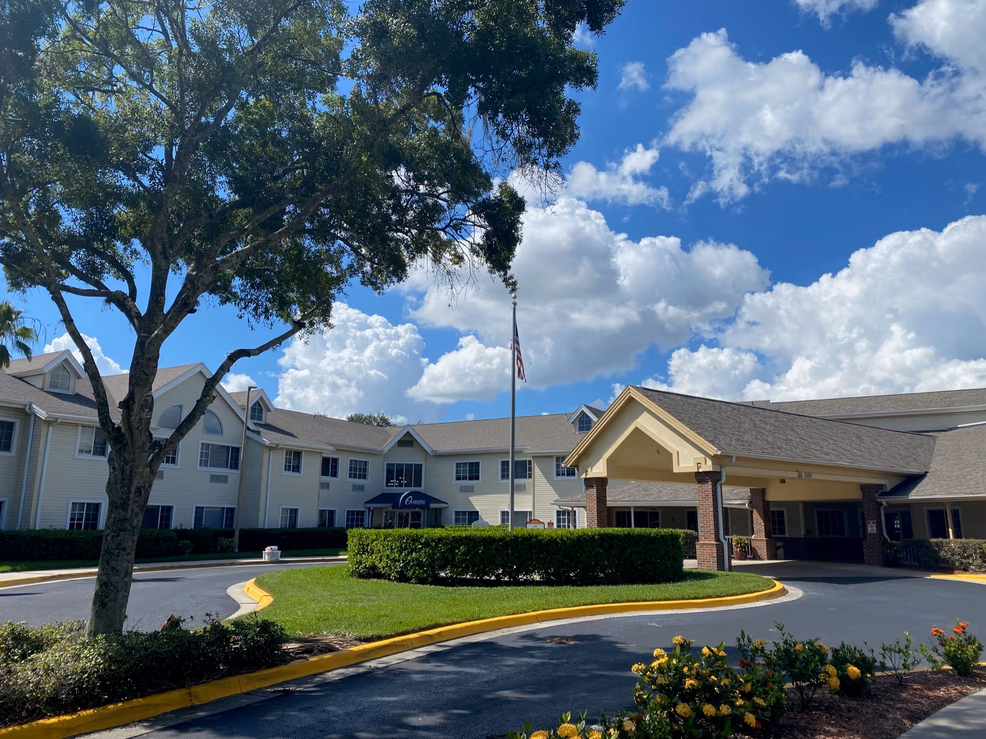Exterior view of a senior living facility named Bridgewalk on Harden, showing a large two-story building with beige siding and a covered entrance. There is a circular driveway with a landscaped island in the center, featuring green bushes and a flagpole with an American flag. The sky is blue with scattered clouds, and there are trees and flowering plants around the driveway.