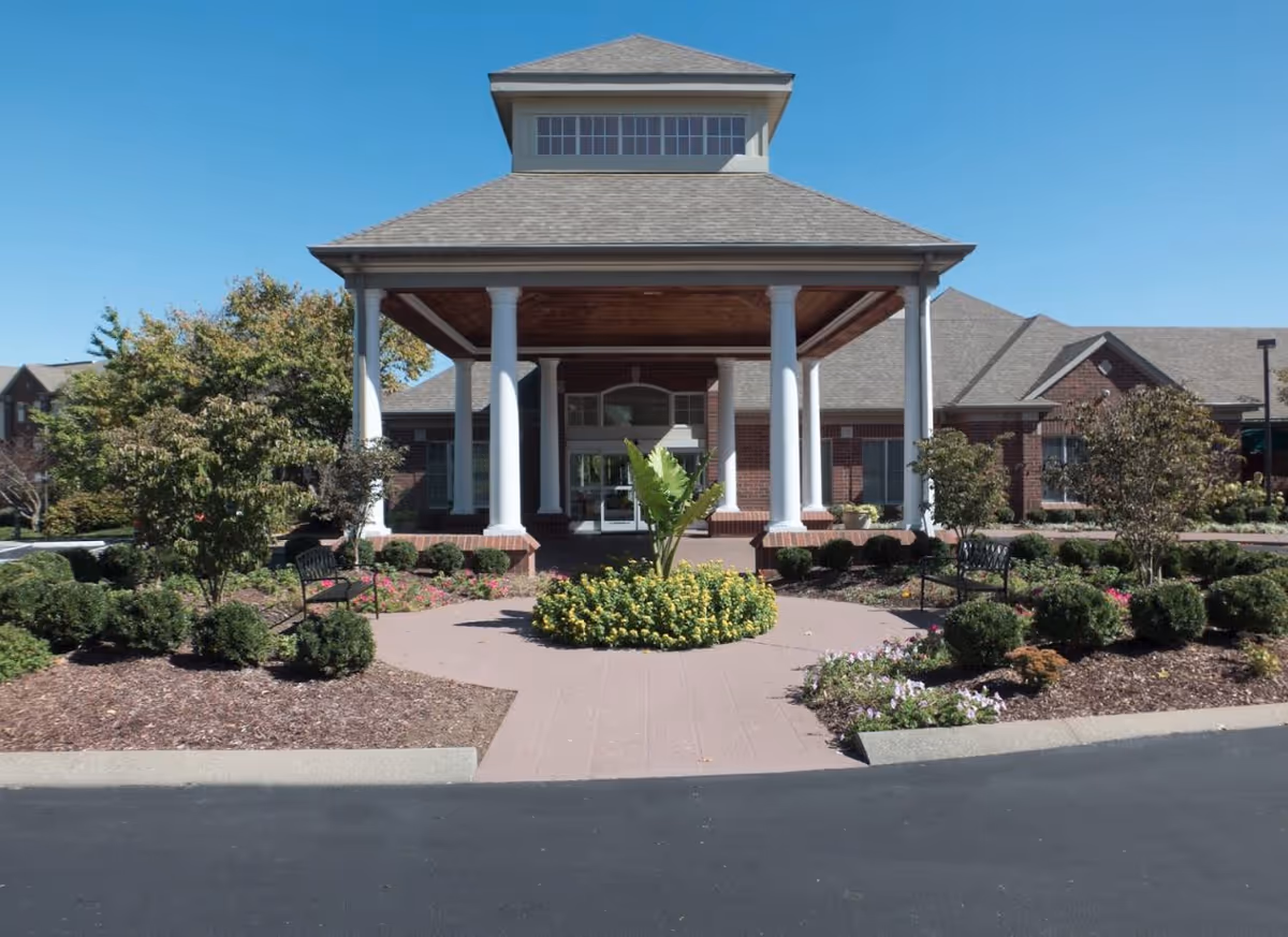 Front entrance of a senior living facility with a covered portico supported by white columns, surrounded by landscaped gardens with bushes, flowers, and benches under a clear blue sky.