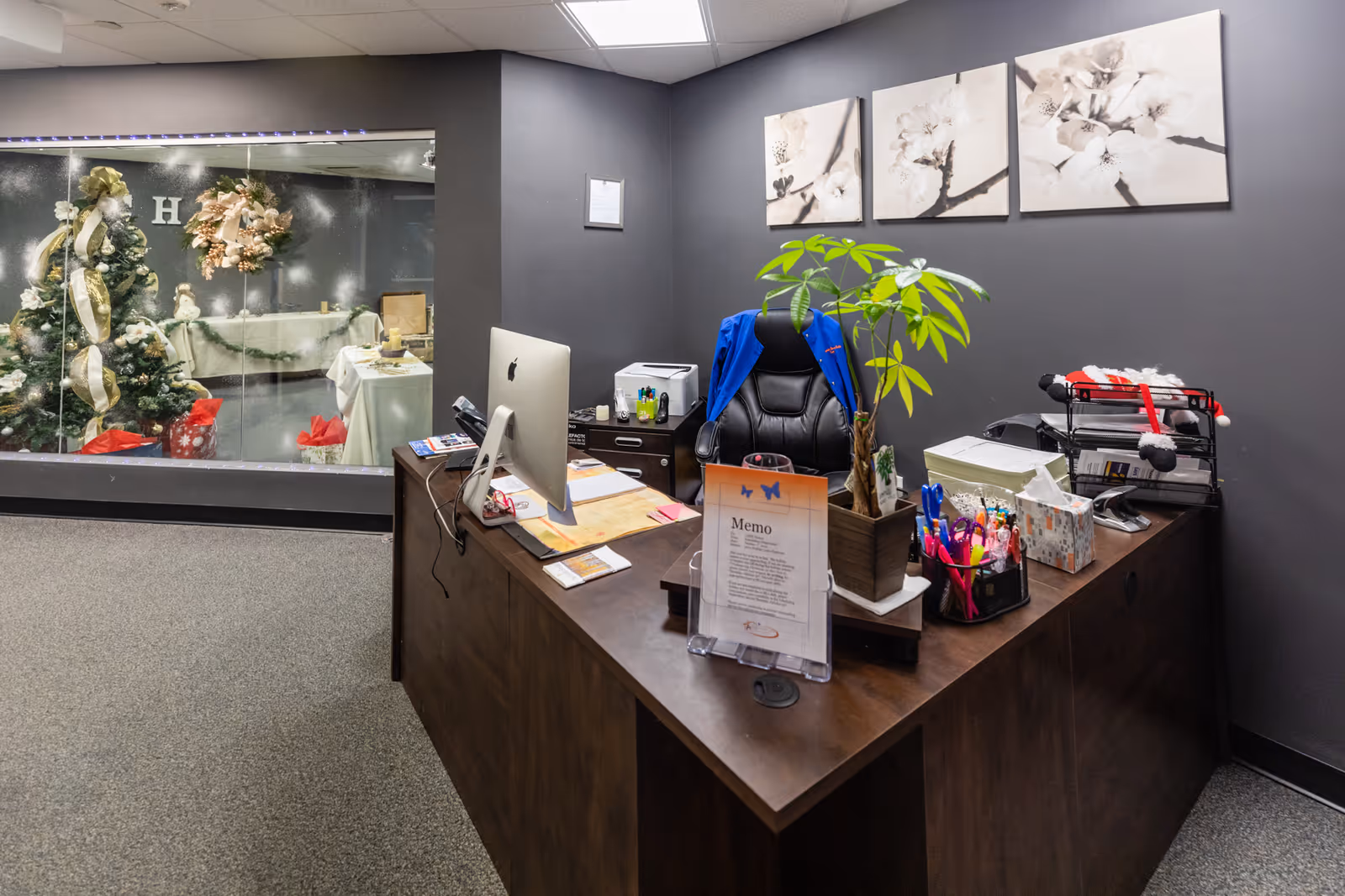 Reception desk in a gray office with plant, computer, and festive decor visible through glass wall.