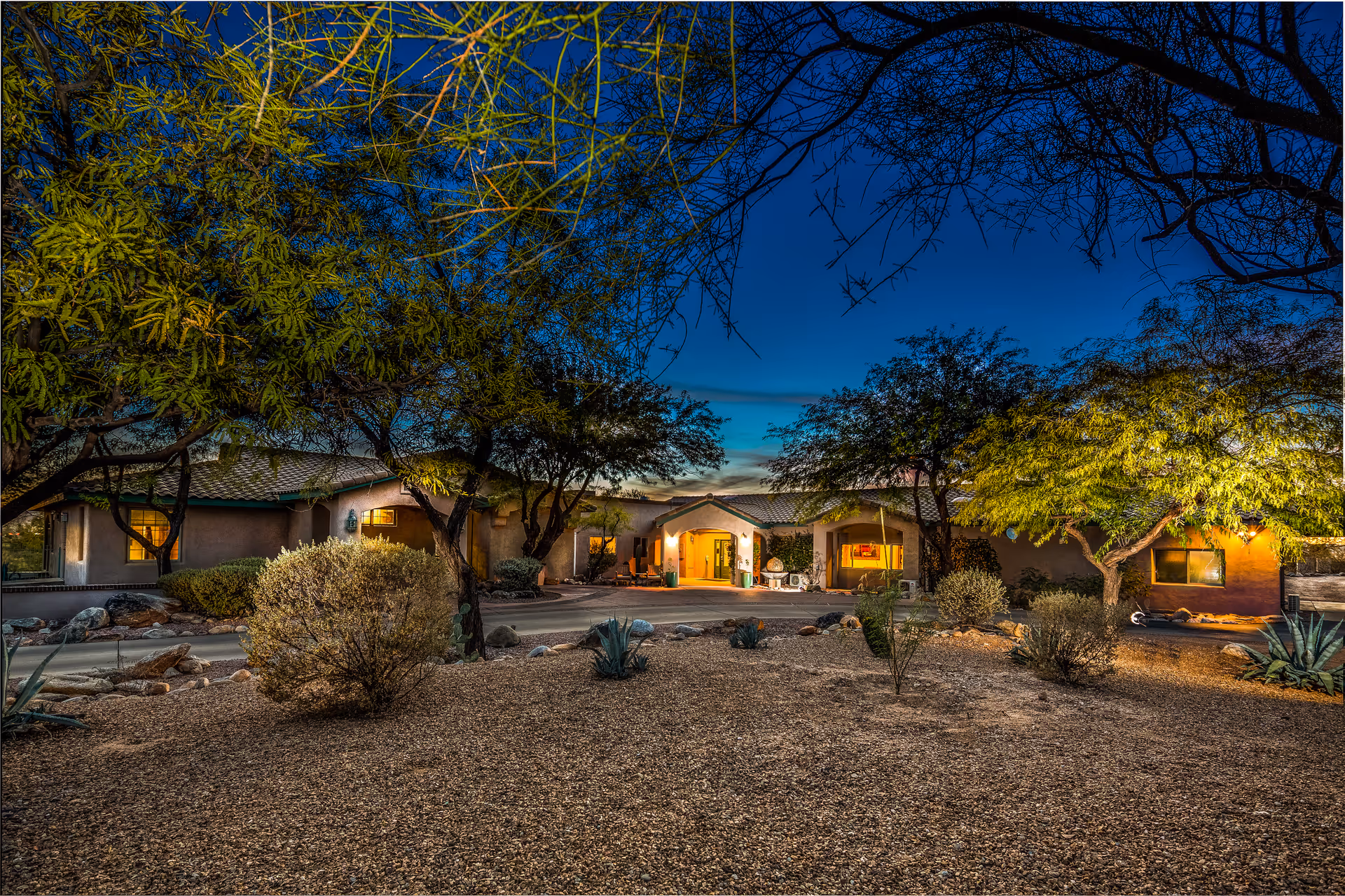 Exterior view of a Mediterranean-style senior living facility at dusk, with desert landscaping and warm lights.