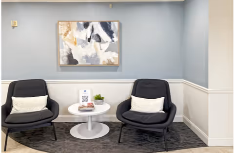 Two dark lounge chairs with white pillows flank a round table in a blue-walled lobby.