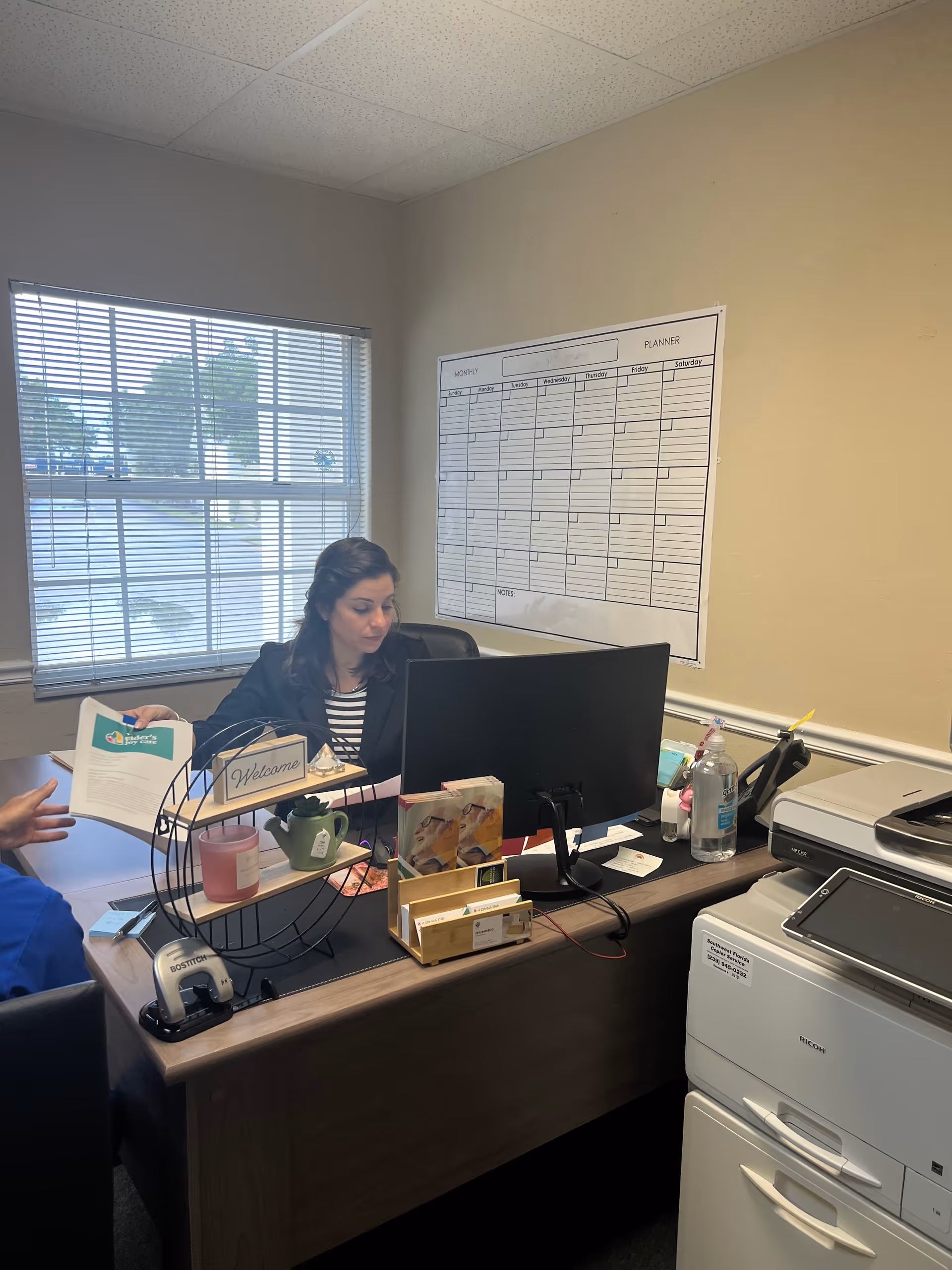 Staff member works at desk in an office with computer, printer, and planner on wall.