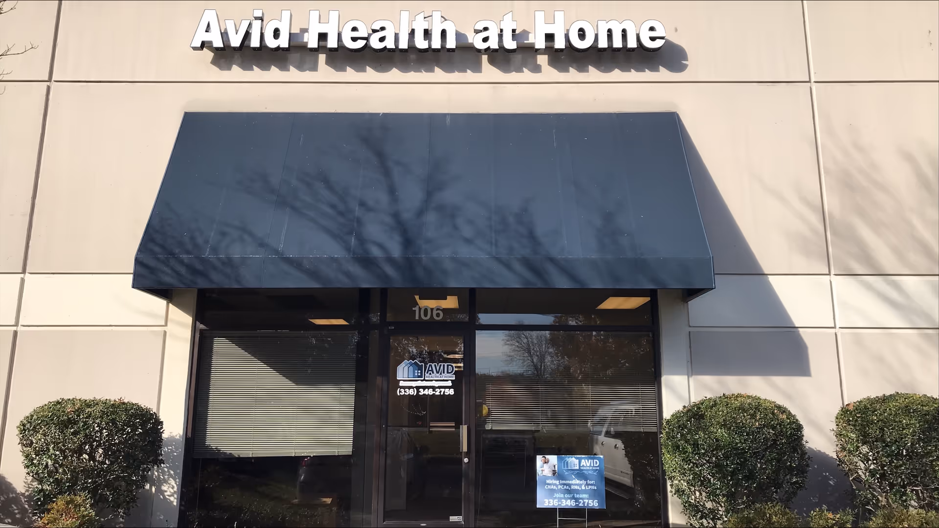 Avid Health at Home storefront with dark glass doors, blue awning, and trimmed shrubs