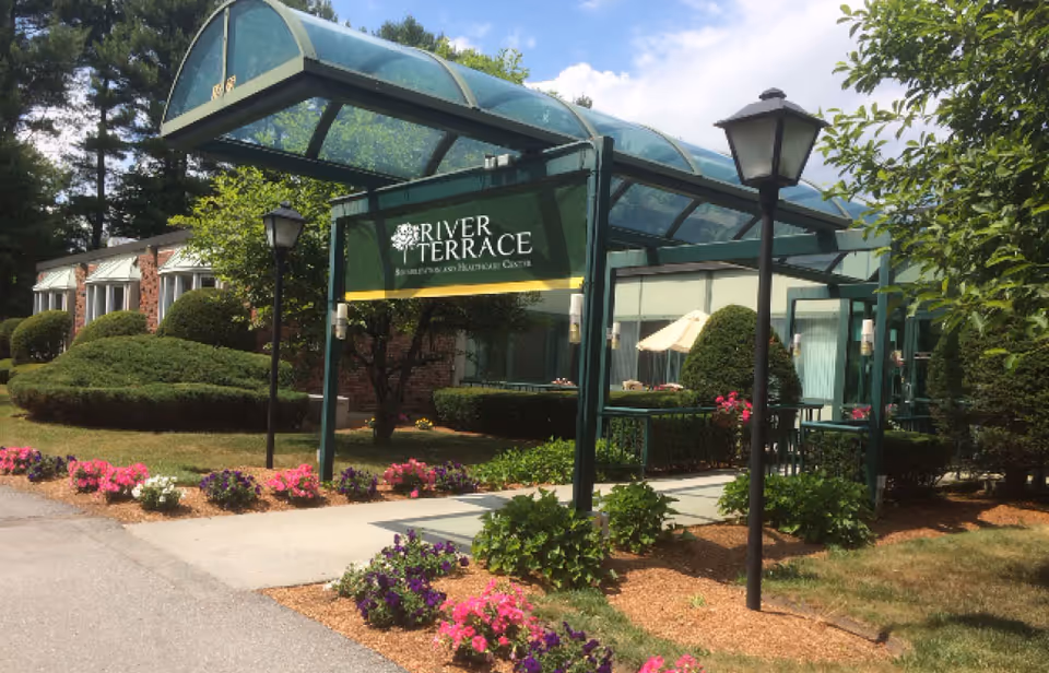 The landscaped front entrance of River Terrace Rehabilitation & Healthcare Center with a green canopy sign, walkway, lamp posts, and flower beds.