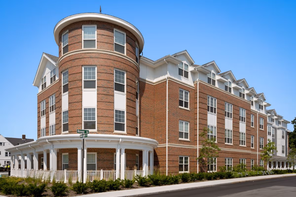 Red-brick, multi-story senior living building with a rounded corner tower and wraparound porch under a clear blue sky.