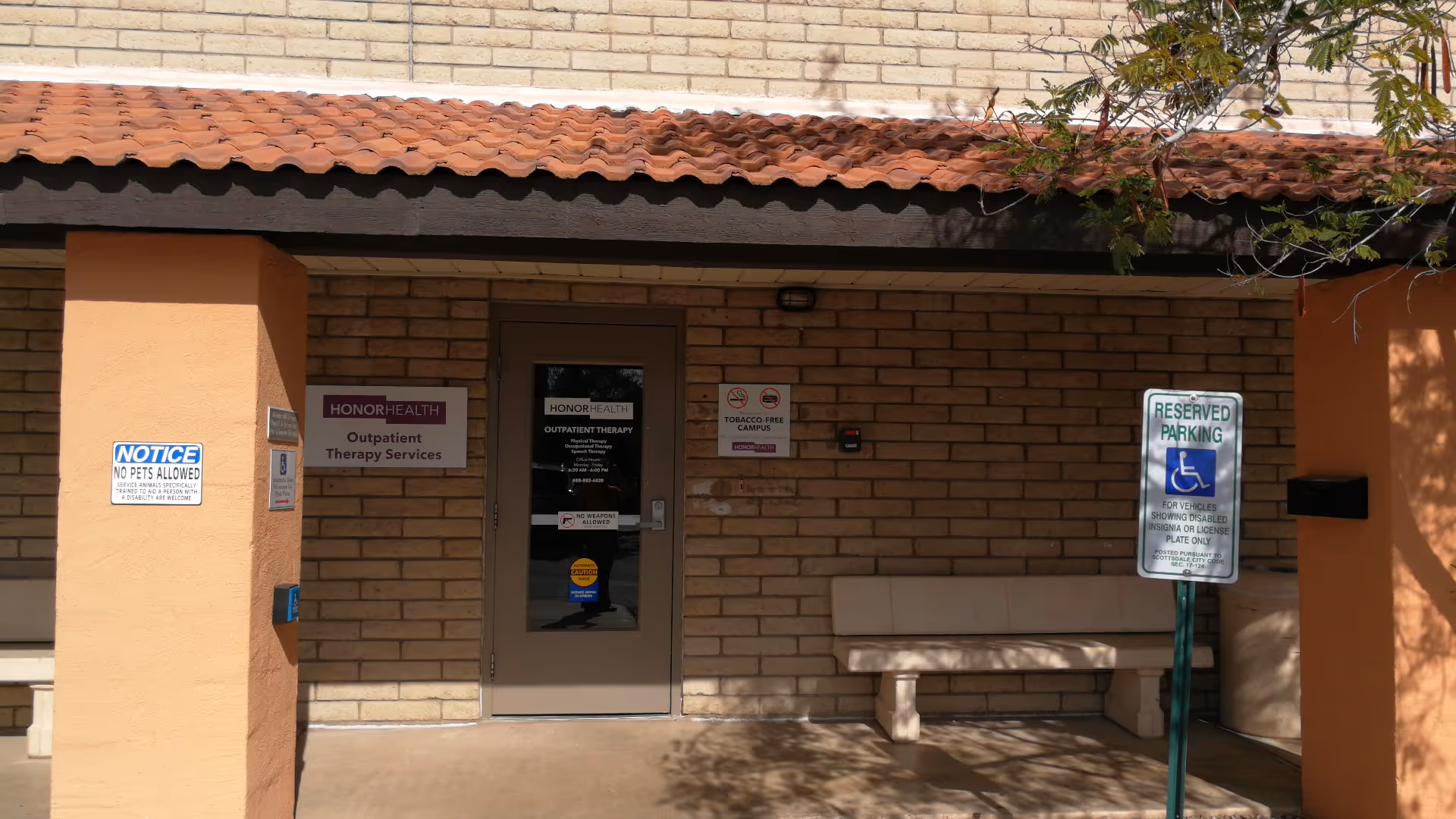 Brick building exterior with HonorHealth door, bench, and handicap parking sign.